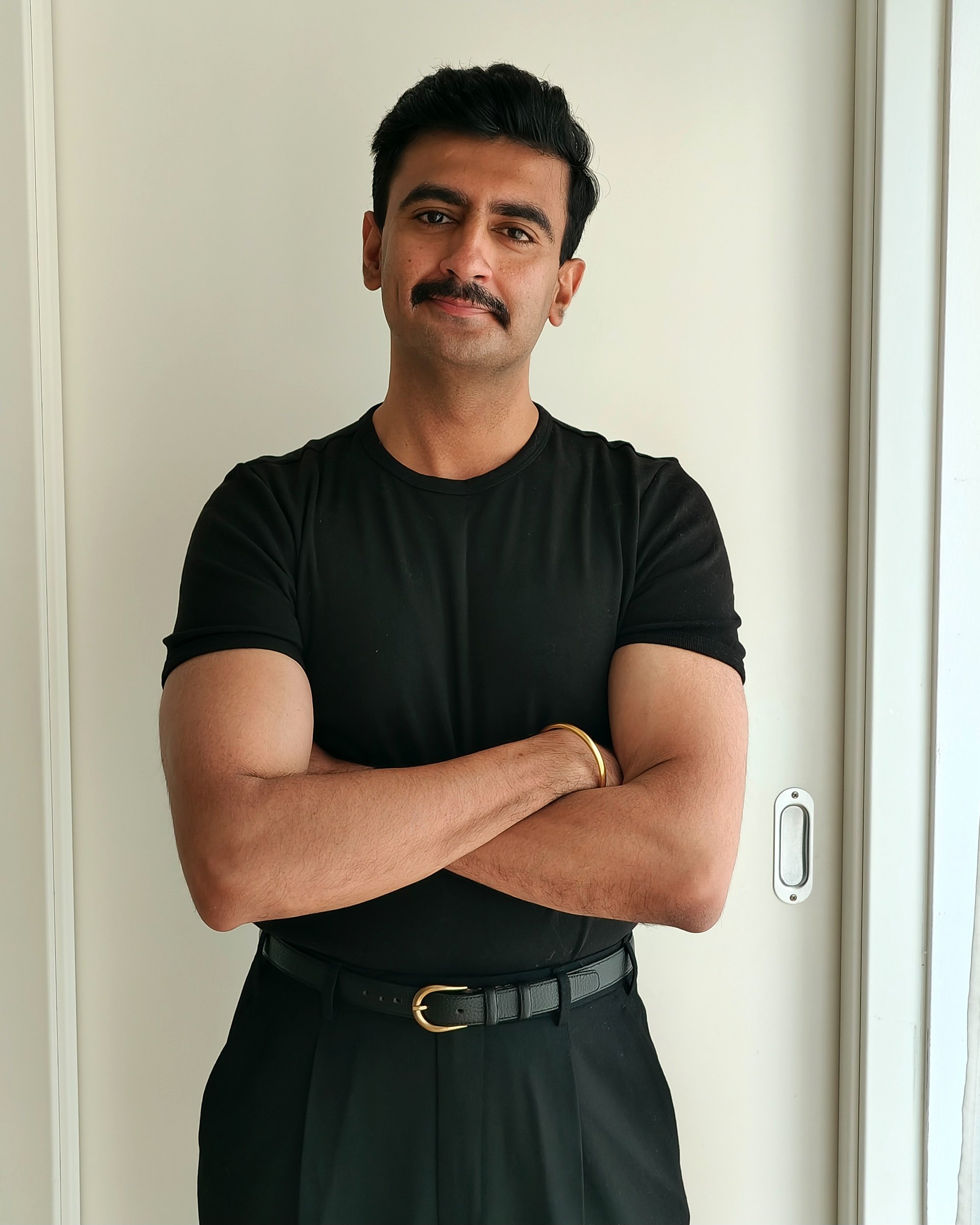 A man with dark hair, a mustache, and a light brown skin tone, wearing a black shirt and a black belt, crossing his arms and standing in front of a white wall and door.