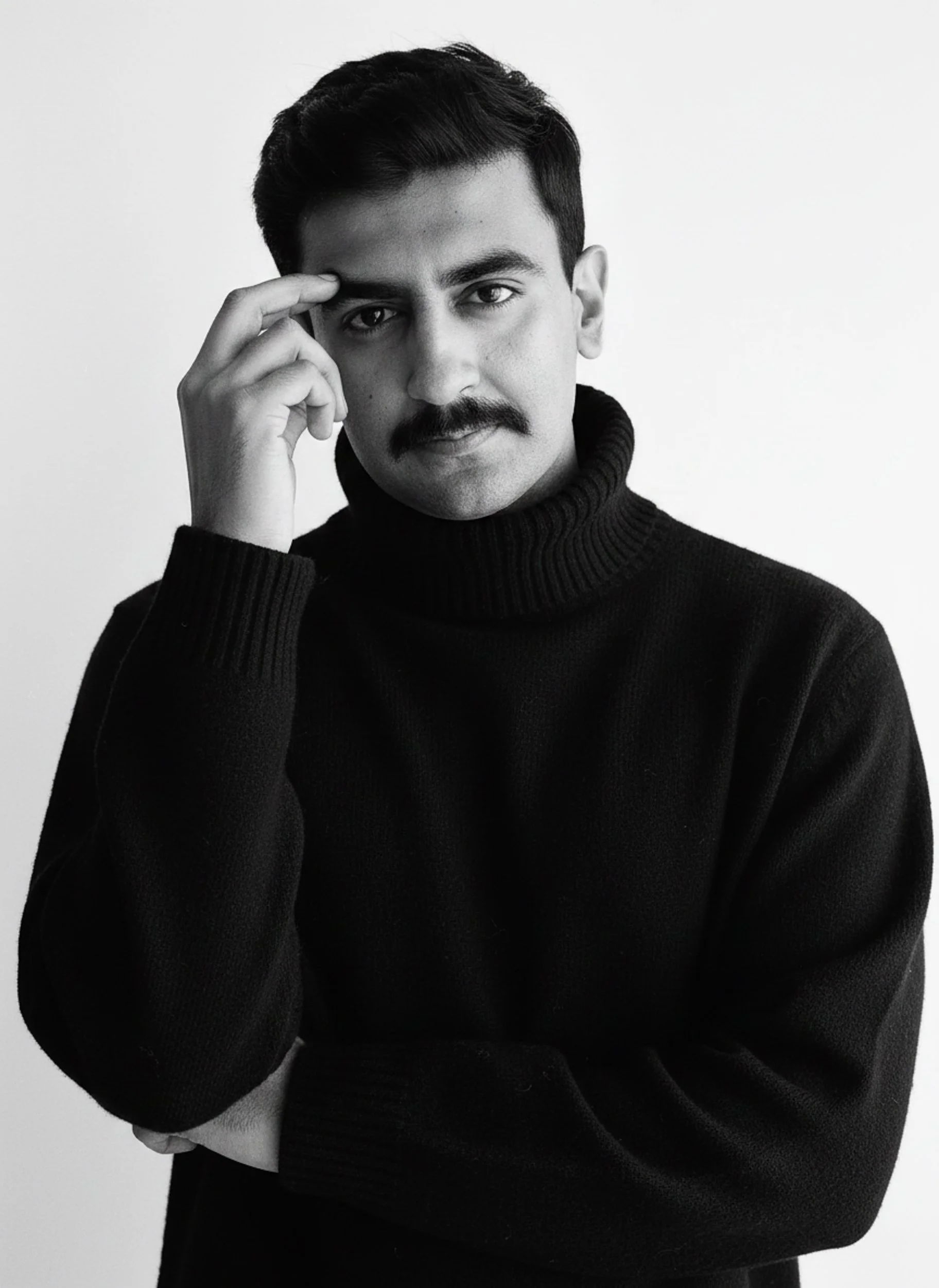 Black and white portrait of a man with a mustache, wearing a turtleneck sweater, touching his forehead with his right hand, against a plain background.