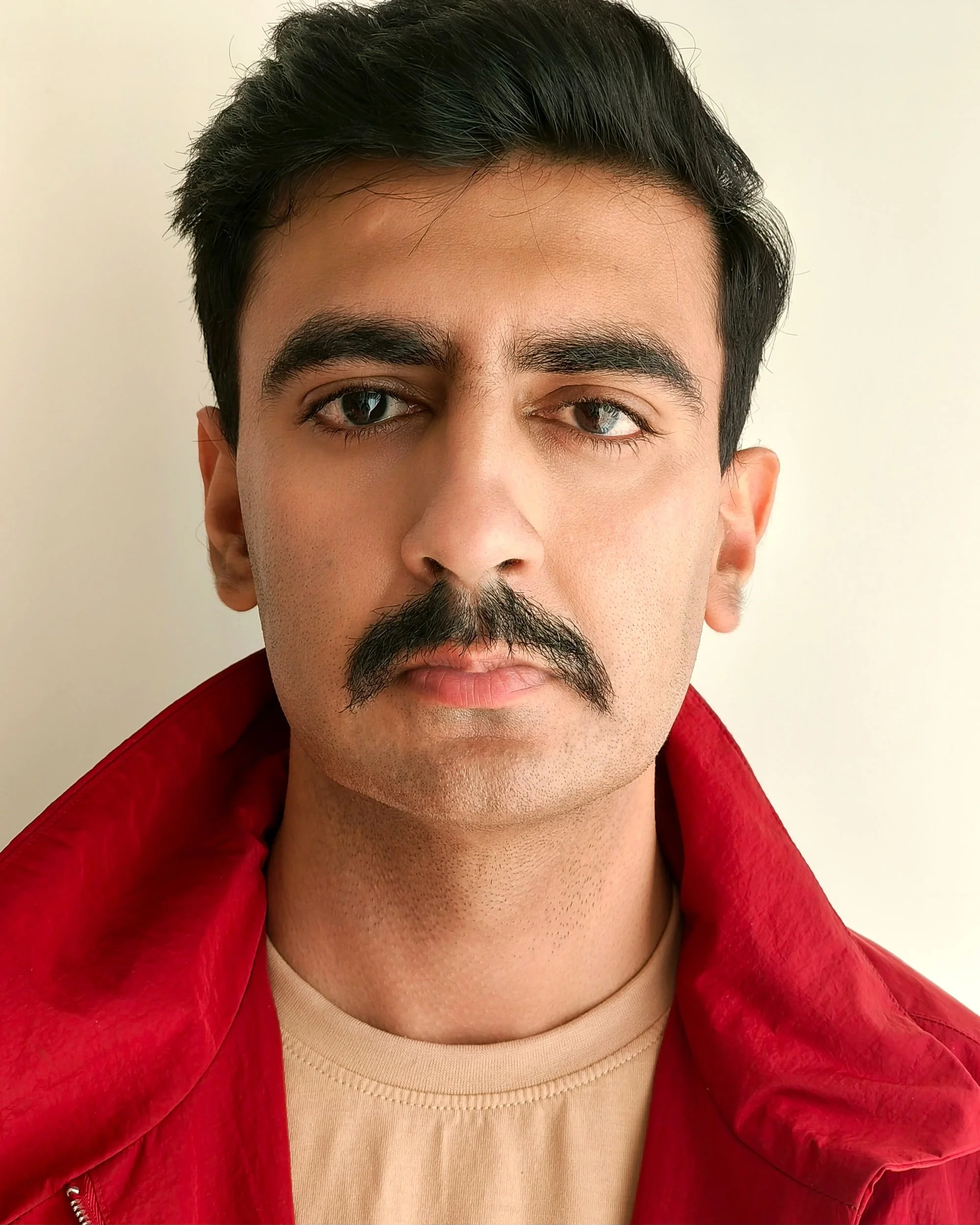 Close-up of a young man with dark hair and a mustache, wearing a beige shirt and a red jacket, standing against a plain white background.