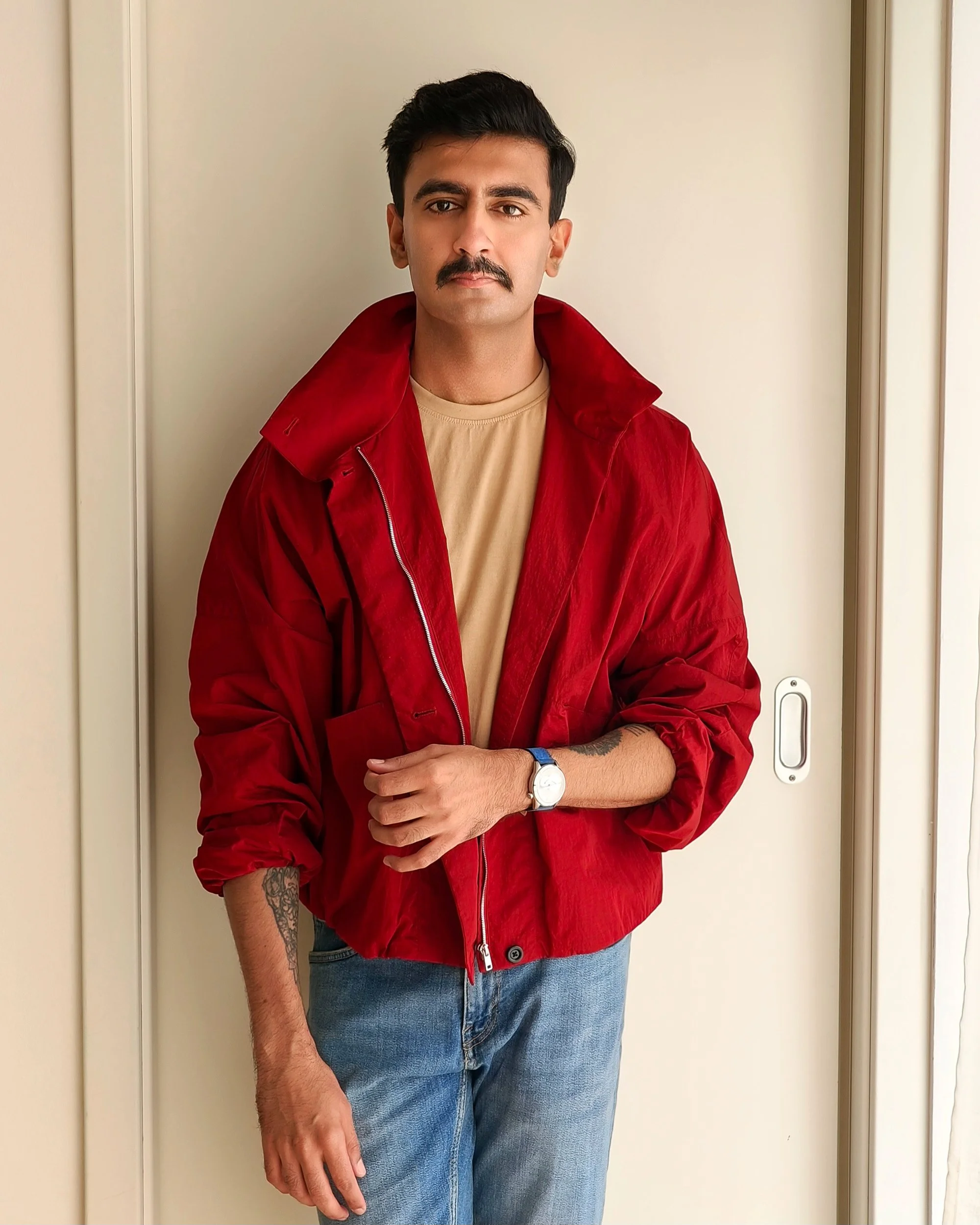 A young man with dark hair and a mustache wearing a beige t-shirt, a red jacket, and blue jeans, standing in front of a plain wall and door.