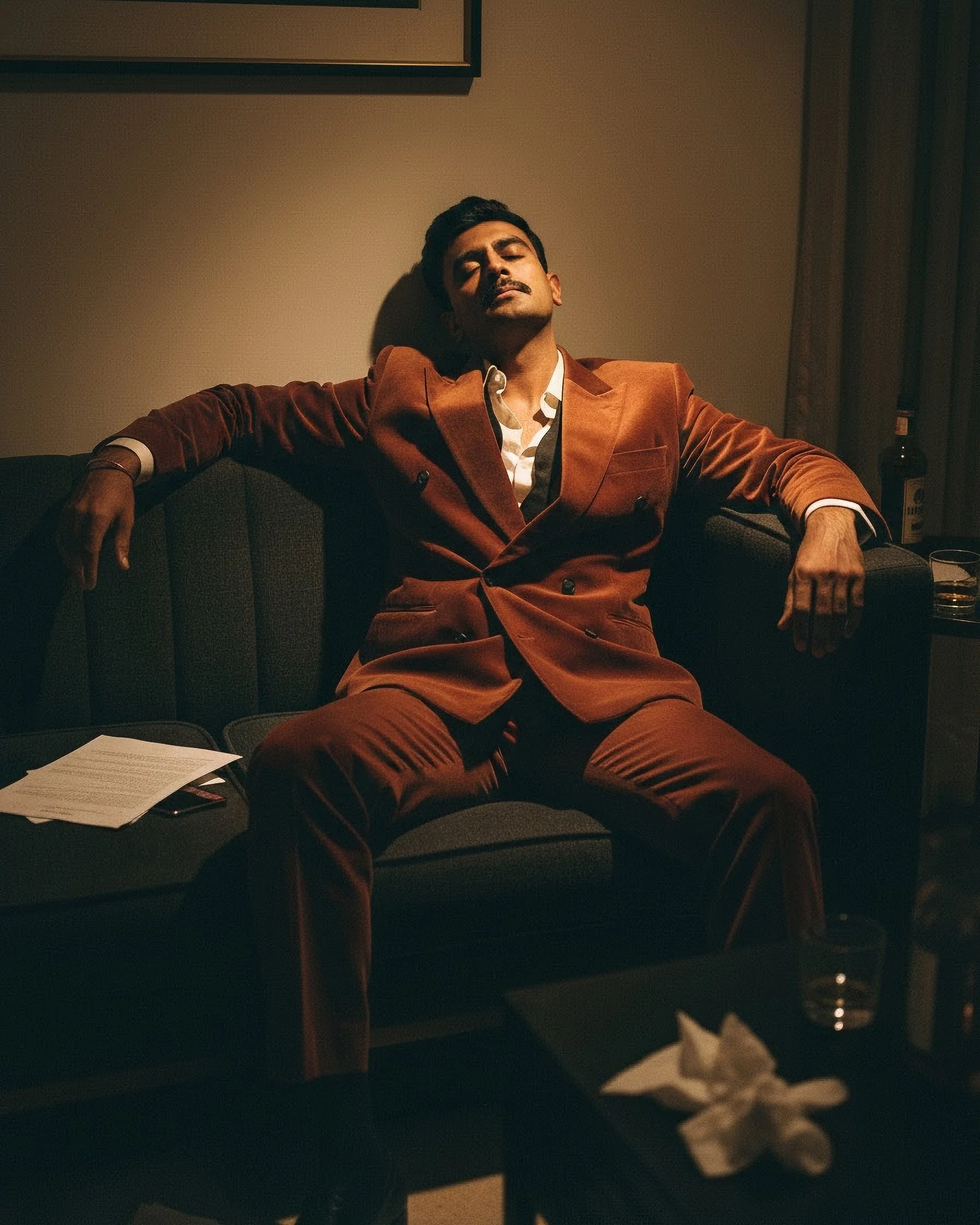 A man wearing a brown suit with a visible white shirt underneath, sitting on a sofa in a dimly lit room with closed curtains, with a table in front holding some papers, and a bottle on a shelf in the background.
