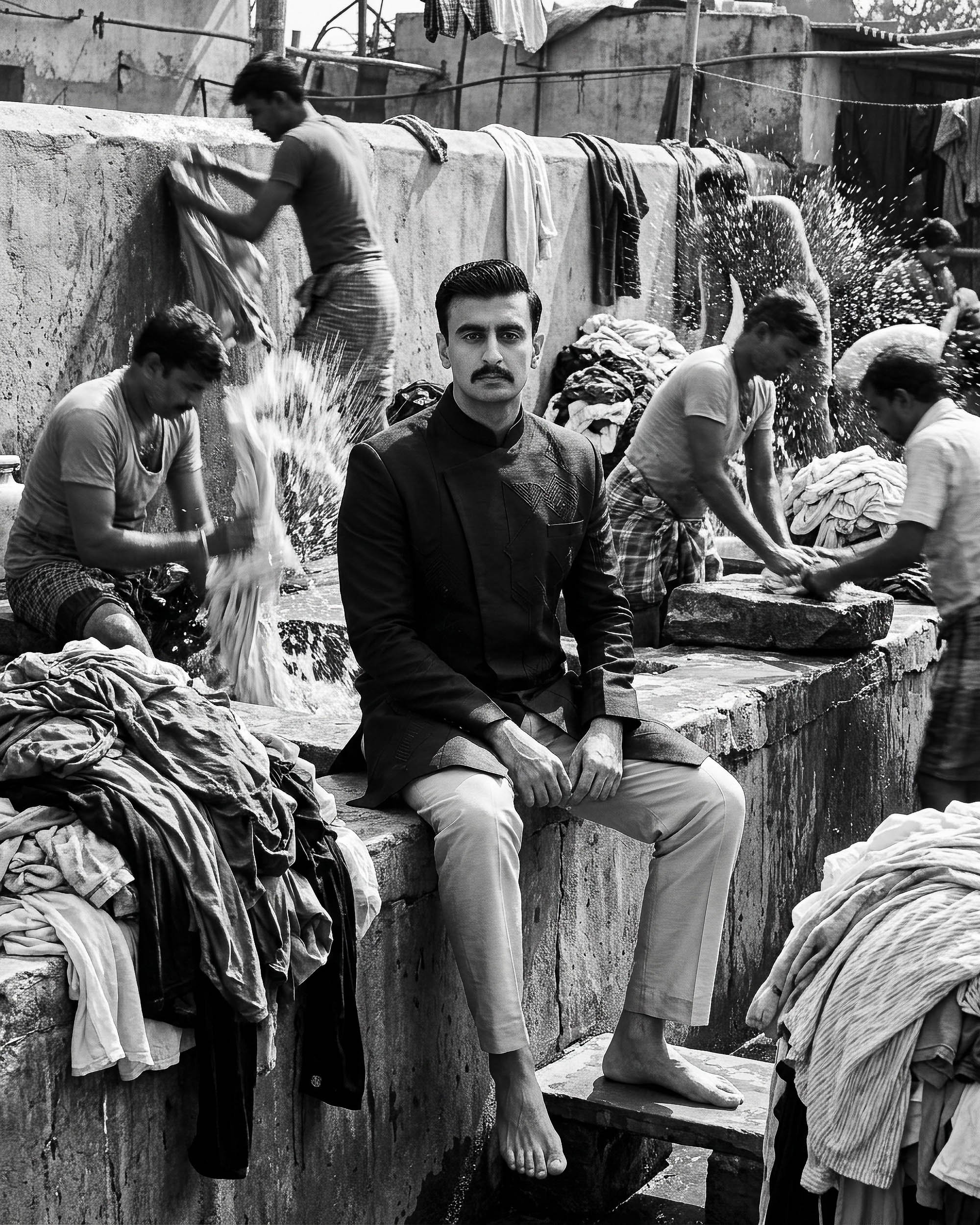 A black and white photo of a man sitting in front of a series of Hindu statues. The statues depict deities, with detailed carvings, and are located outdoors. The man has dark hair, a mustache, and is wearing traditional Indian attire, looking directl