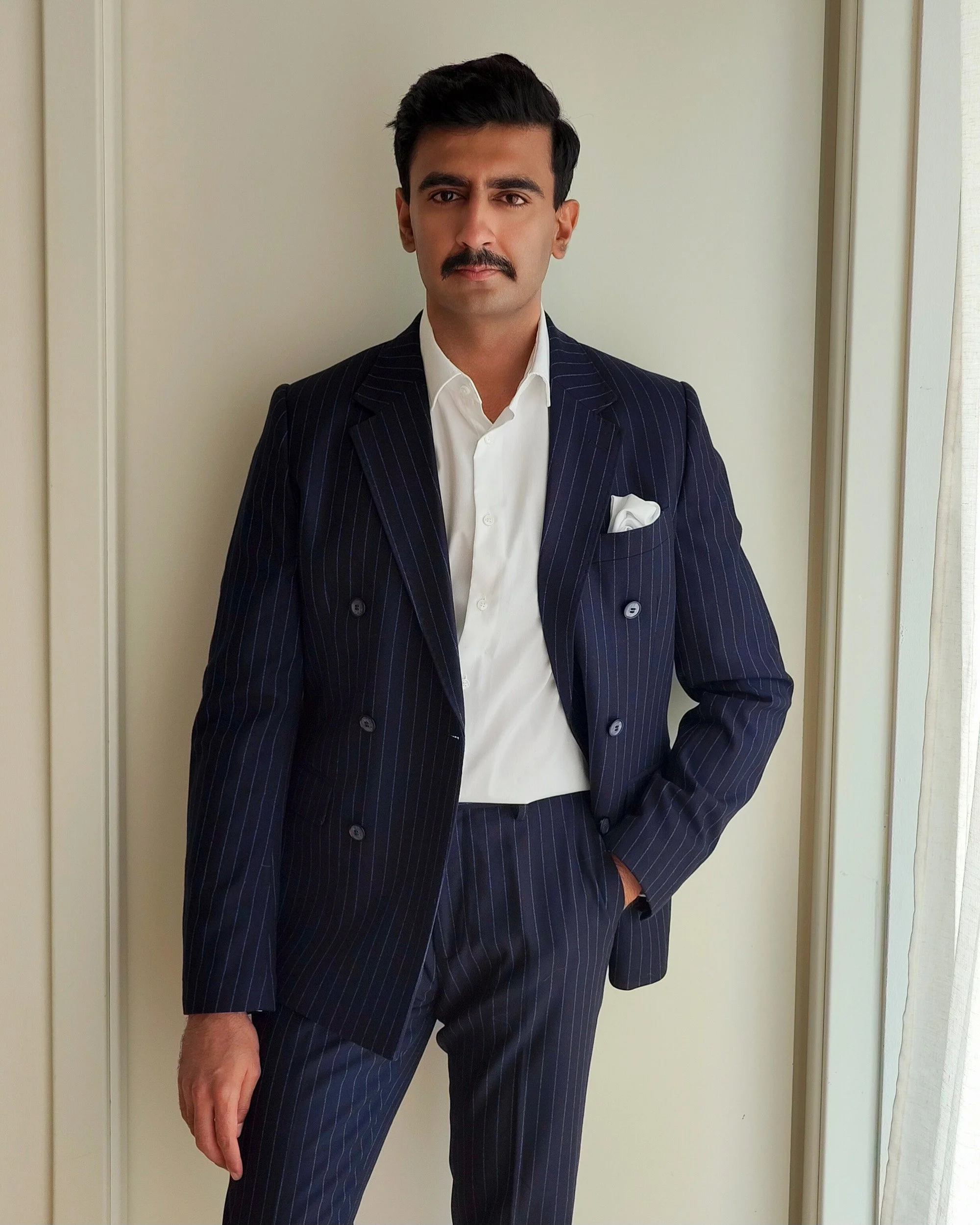 A well-dressed man in a dark pinstripe suit with a white shirt and a pocket square, standing near a door and posing confidently.