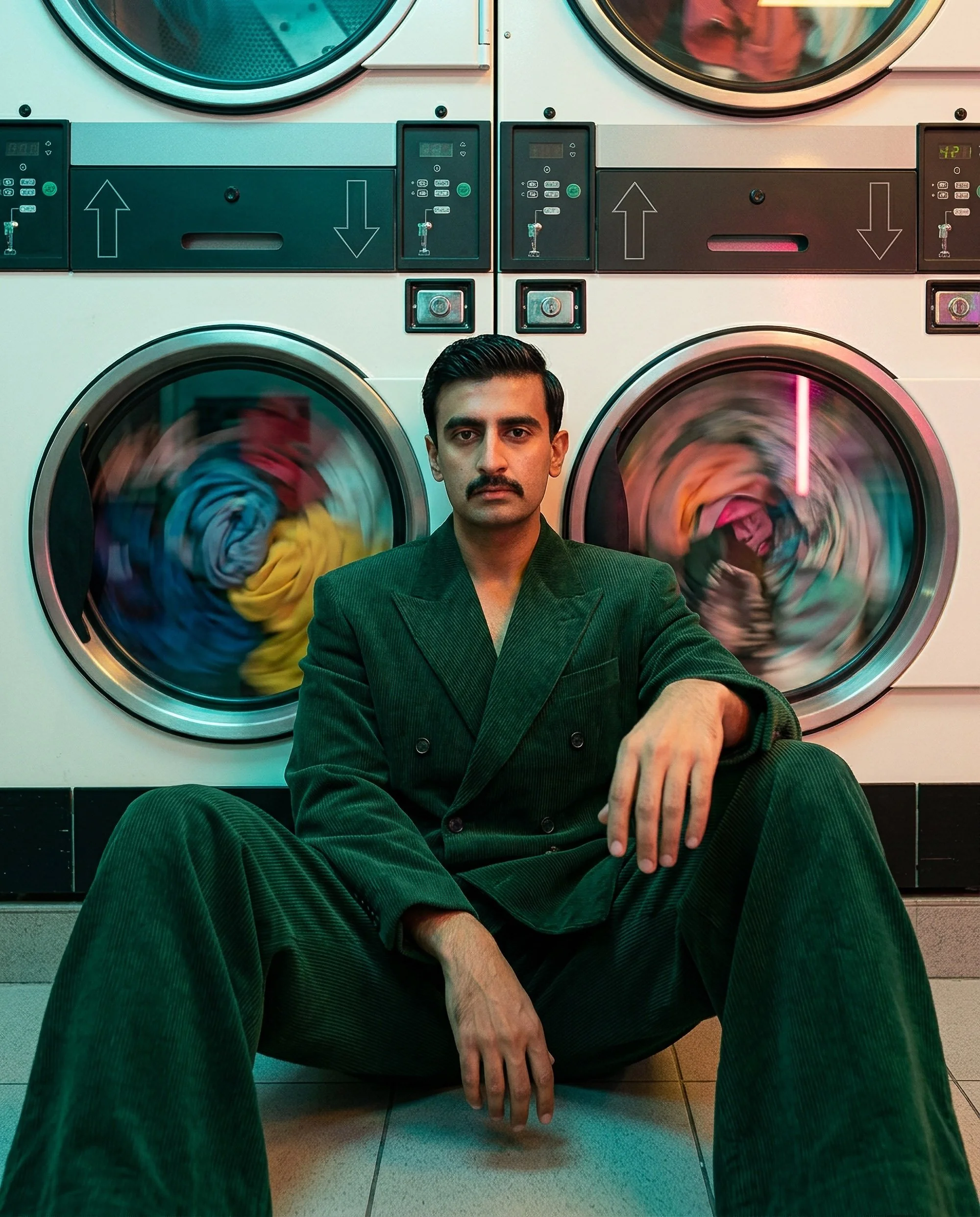A man with a mustache wearing a dark green, pinstripe suit sitting in front of a row of washing machines in a laundromat. The washing machines are side by side, with one on the left showing laundry in motion and a second on the right with laundry spi