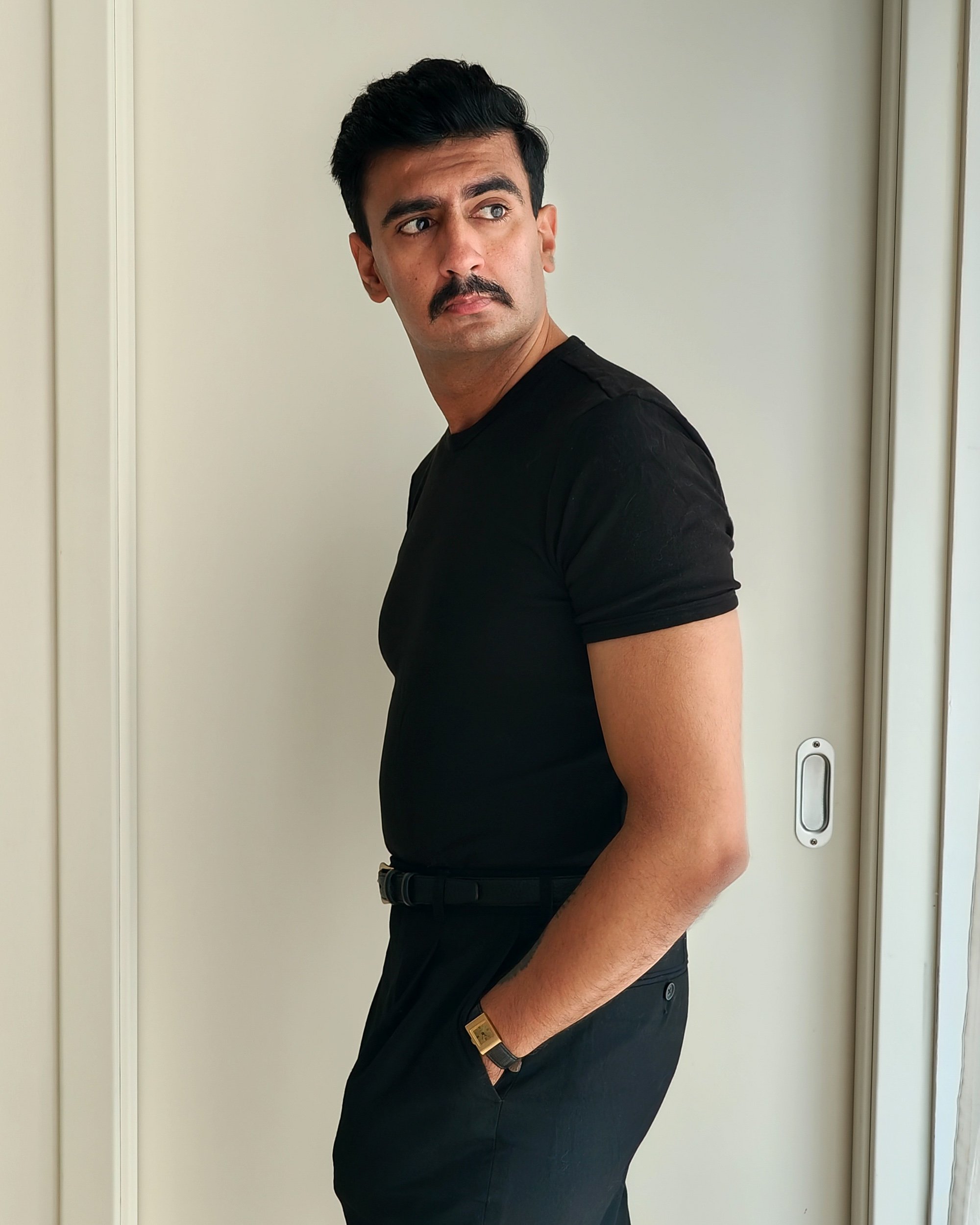 A man with dark hair and a mustache wearing a black shirt and watch, standing sideways with hands in pockets, near a white door.