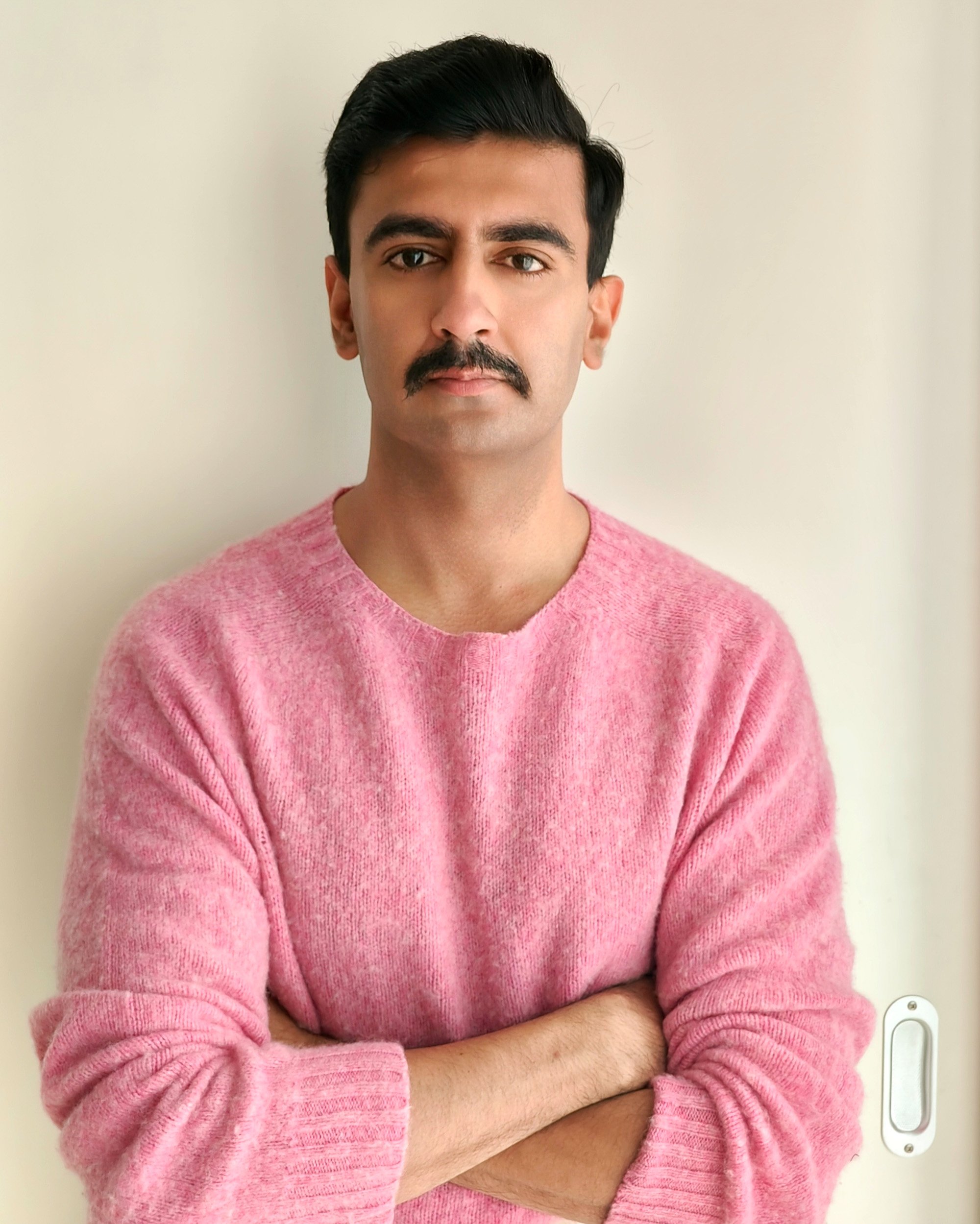 A person wearing a pink sweater with arms crossed, standing against a plain light-colored wall.