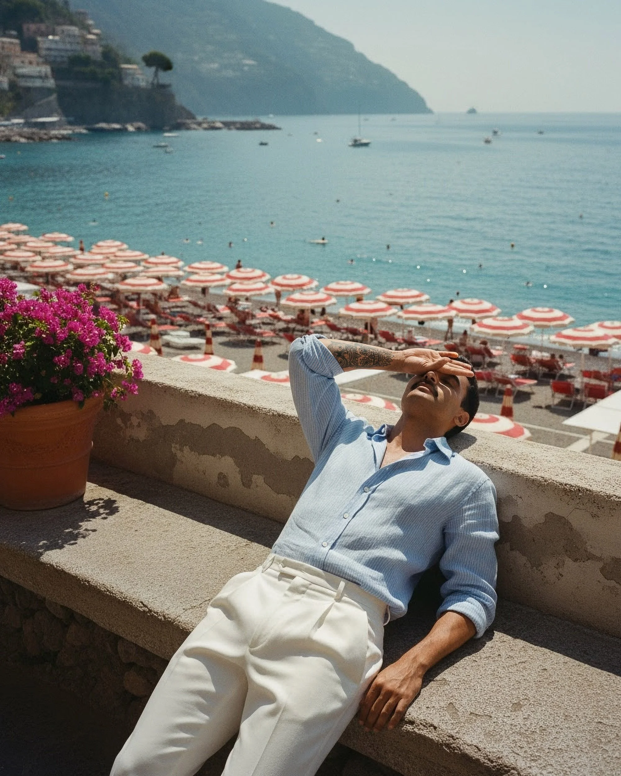 A man with tattoos on his arm is lounging on a concrete ledge, shielding his eyes from the sun with his hand. Behind him, there are rows of red and white striped umbrellas and a view of the ocean with boats, near a hillside town.