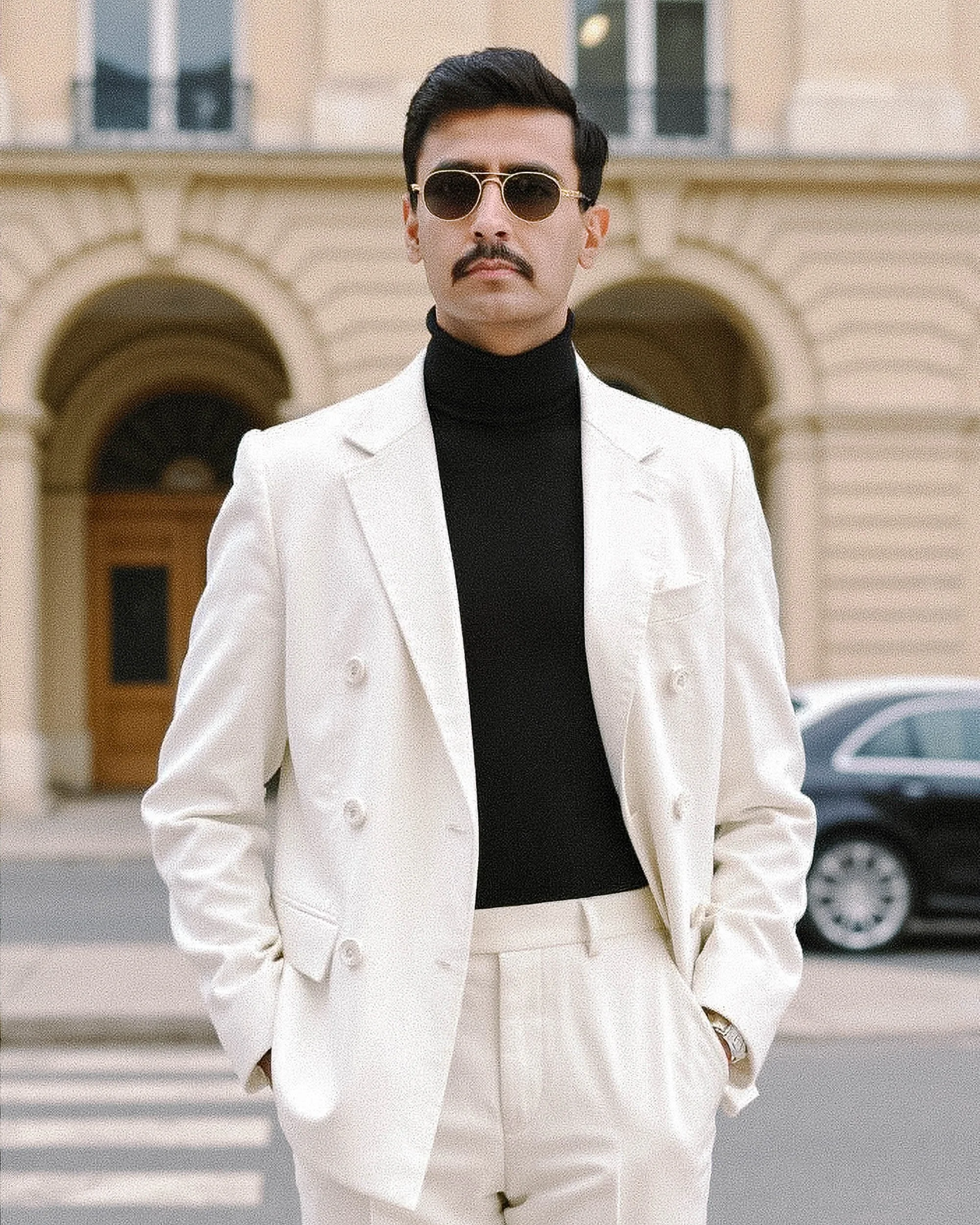 A man with dark hair, a mustache, and sunglasses, wearing a white blazer over a black turtleneck, standing outdoors in front of a beige building with arches.