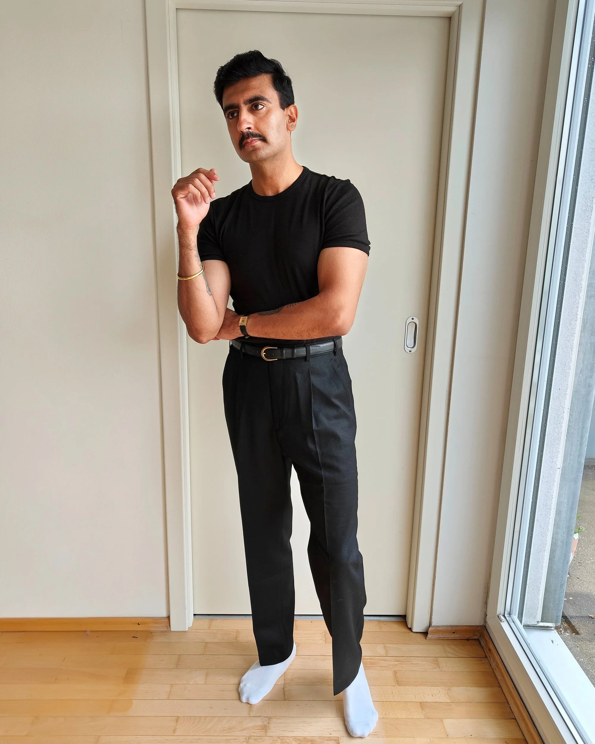 A man with dark hair and a mustache dressed in a black t-shirt and dark pants, standing indoors near a sliding glass door, with one hand near his face and the other crossed over his chest, wearing a gold bracelet and a watch, and socks but no shoes.