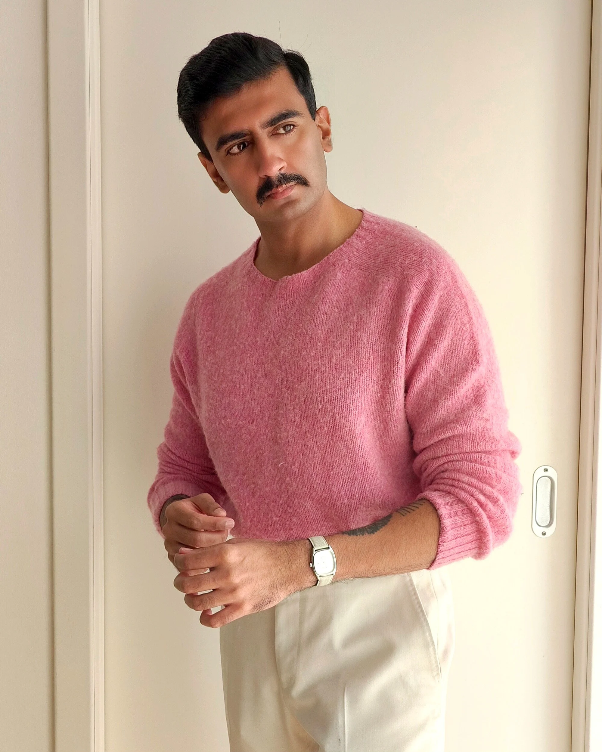 A person with dark hair and a mustache is wearing a pink sweater and light-colored pants, standing indoors near a beige wall and a closed door.