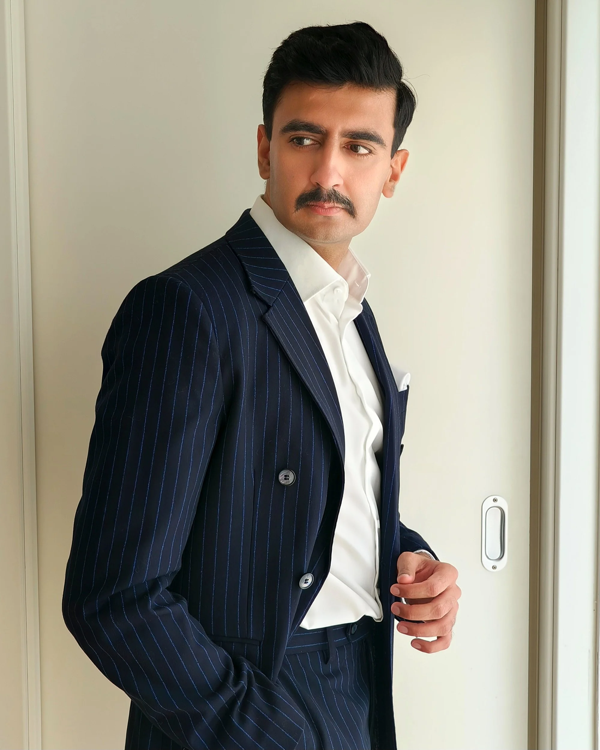 A man in a pinstripe navy suit and white dress shirt standing near a wall, with his hand holding a device, looking to the side.