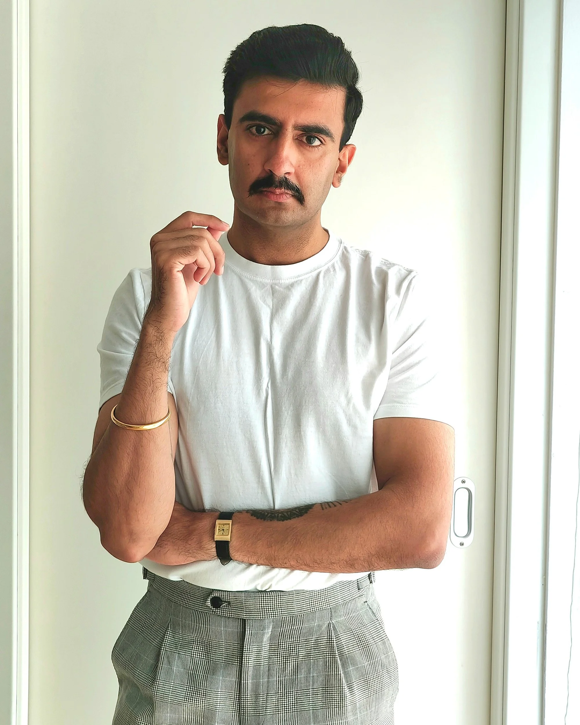 A man with dark hair, a mustache, wearing a white t-shirt, plaid pants, and gold watch, standing with arms crossed in front of a white door.