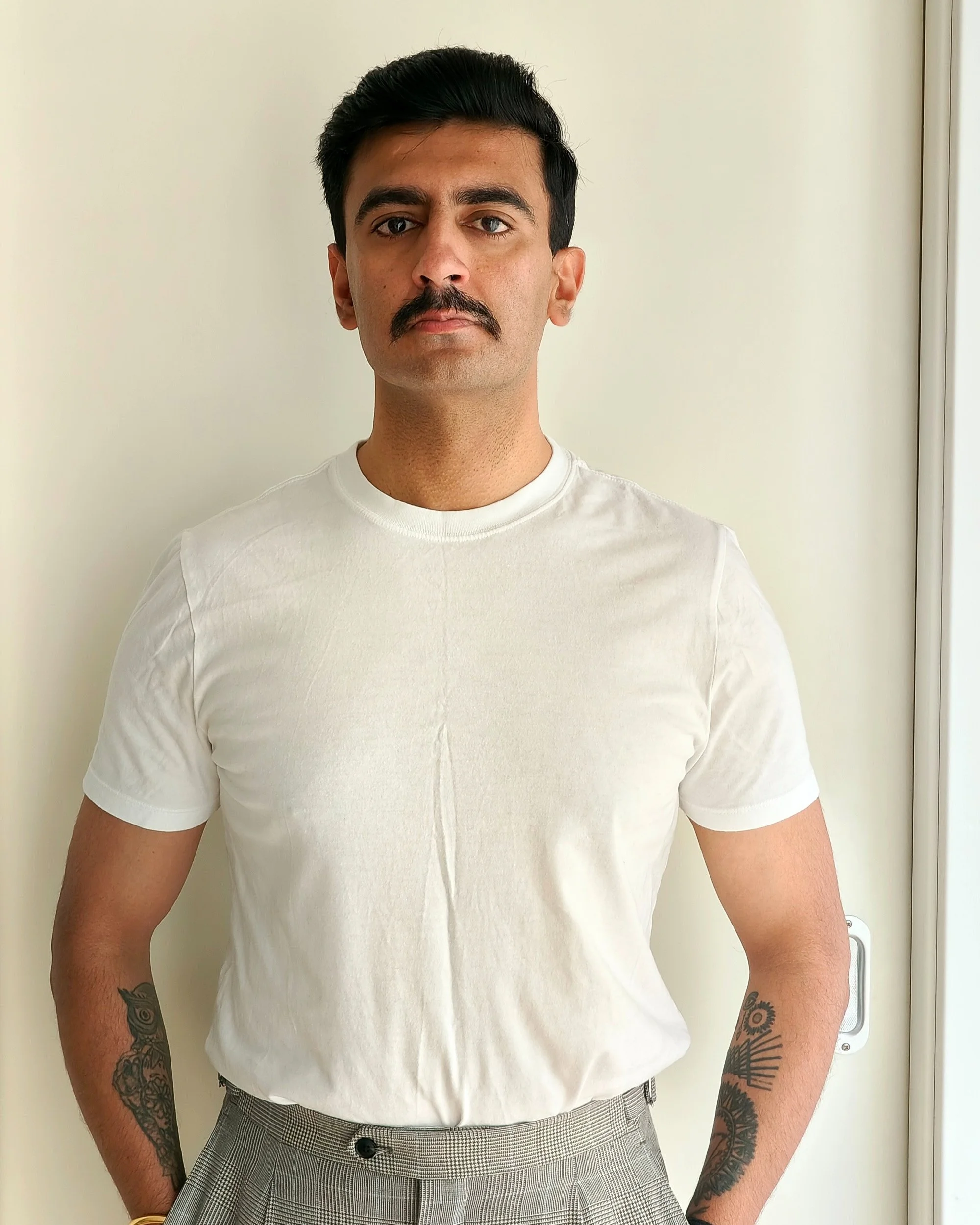 A young man with dark hair, mustache, and tattoos on his arms, wearing a white t-shirt and checkered trousers, standing against a plain white wall.