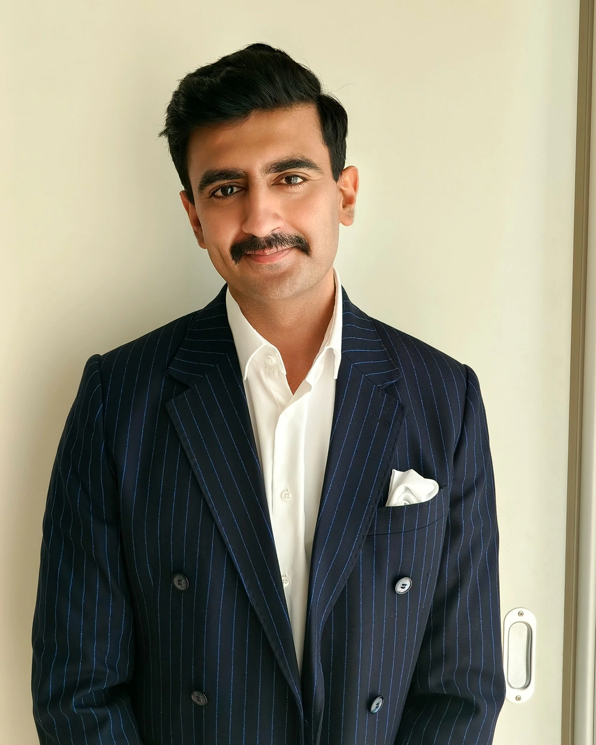 A man with dark hair and a mustache wearing a dark pinstripe suit and white shirt, smiling slightly, against a plain light-colored background.