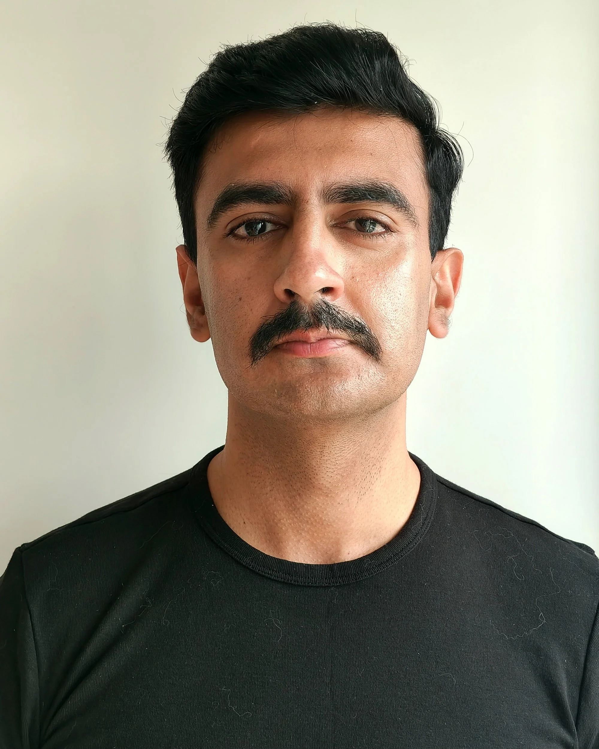 A man with dark hair, a mustache, and beard wearing a black shirt, standing in front of a plain light-colored background.