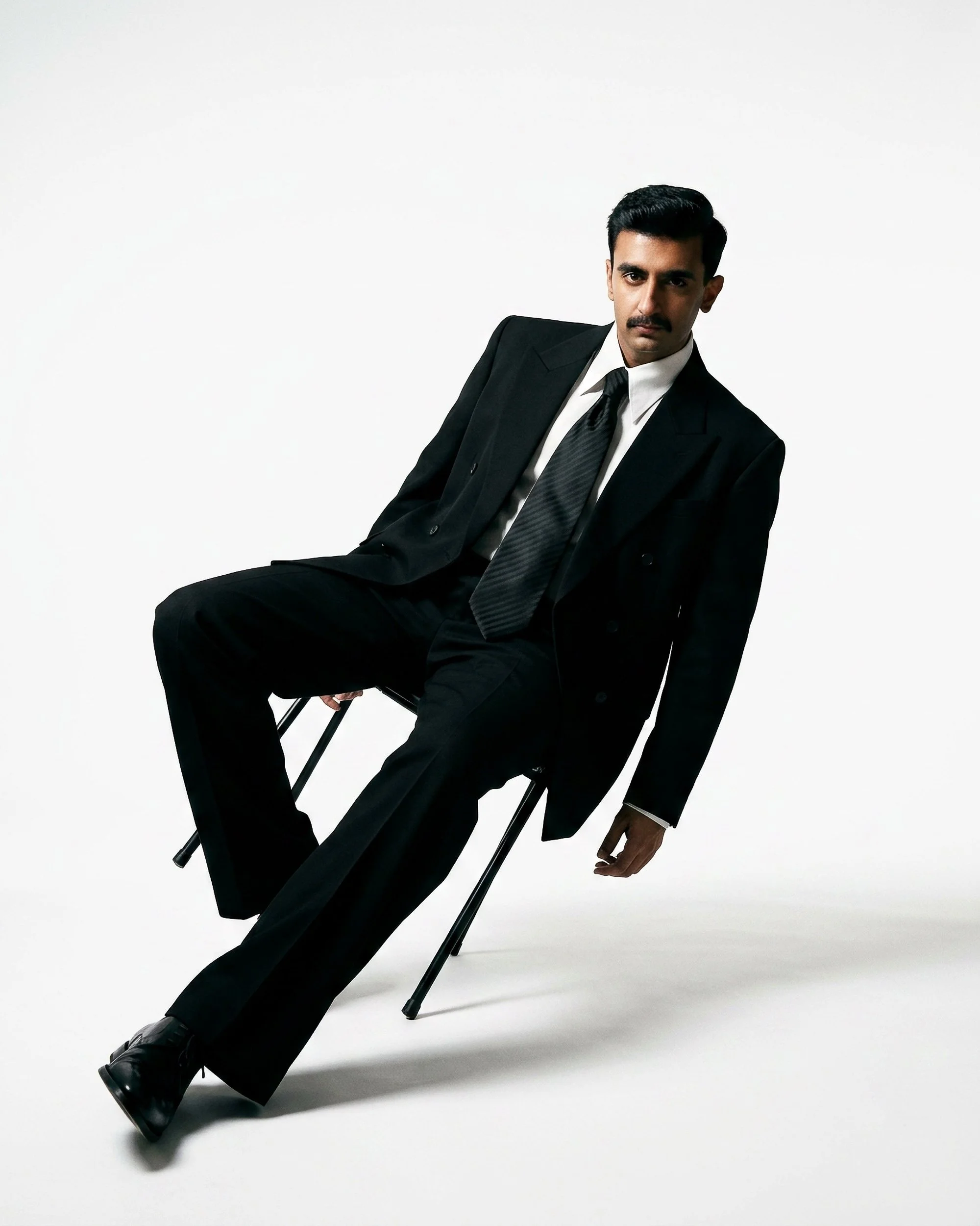 A man with dark hair, mustache, and medium skin tone sitting on a foldable black chair against a white background. He is dressed in a formal black suit, white shirt, and black tie, and looks directly at the camera.
