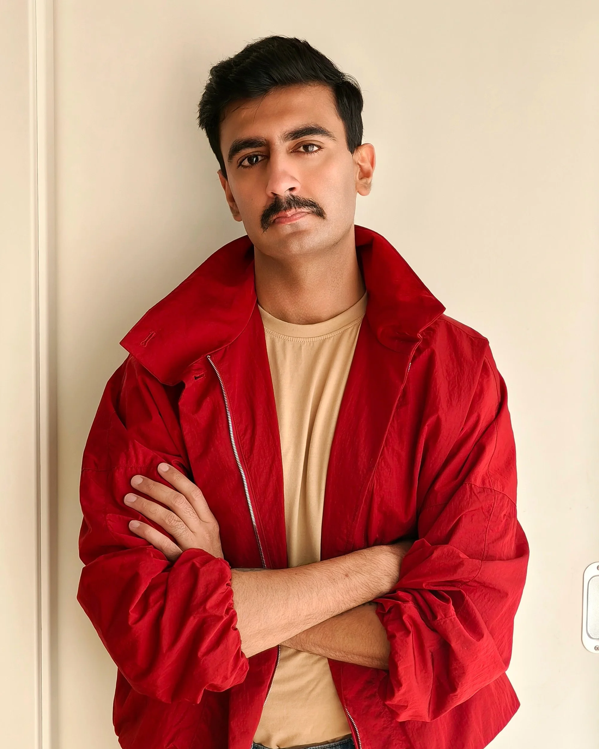 A young man with a mustache and dark hair standing against a plain wall. He is wearing a beige shirt and a red jacket, with his arms crossed and a serious expression on his face.