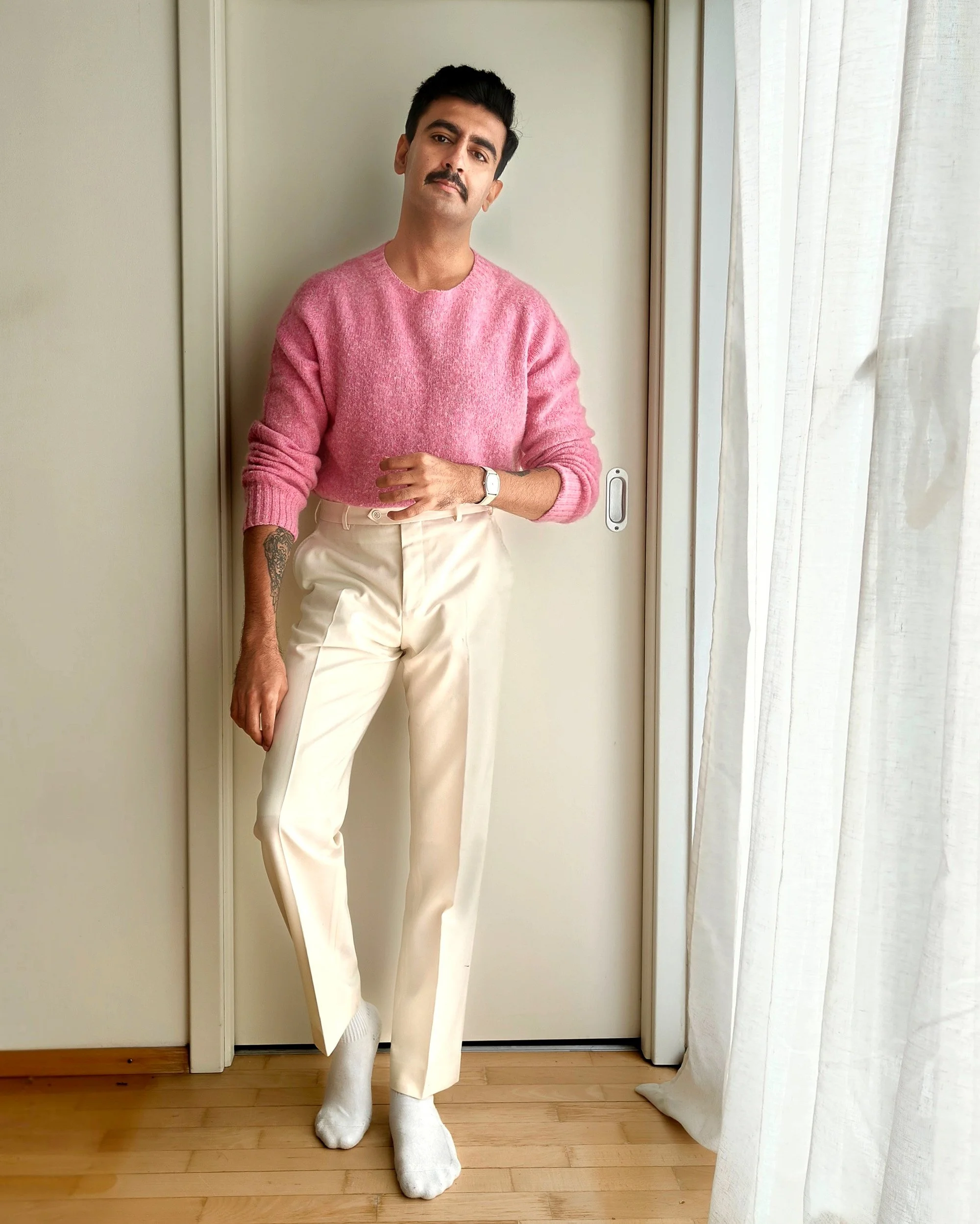A man with dark hair and a mustache wearing a pink sweater and beige pants, standing indoors on a hardwood floor near a sliding door and light-colored curtains.