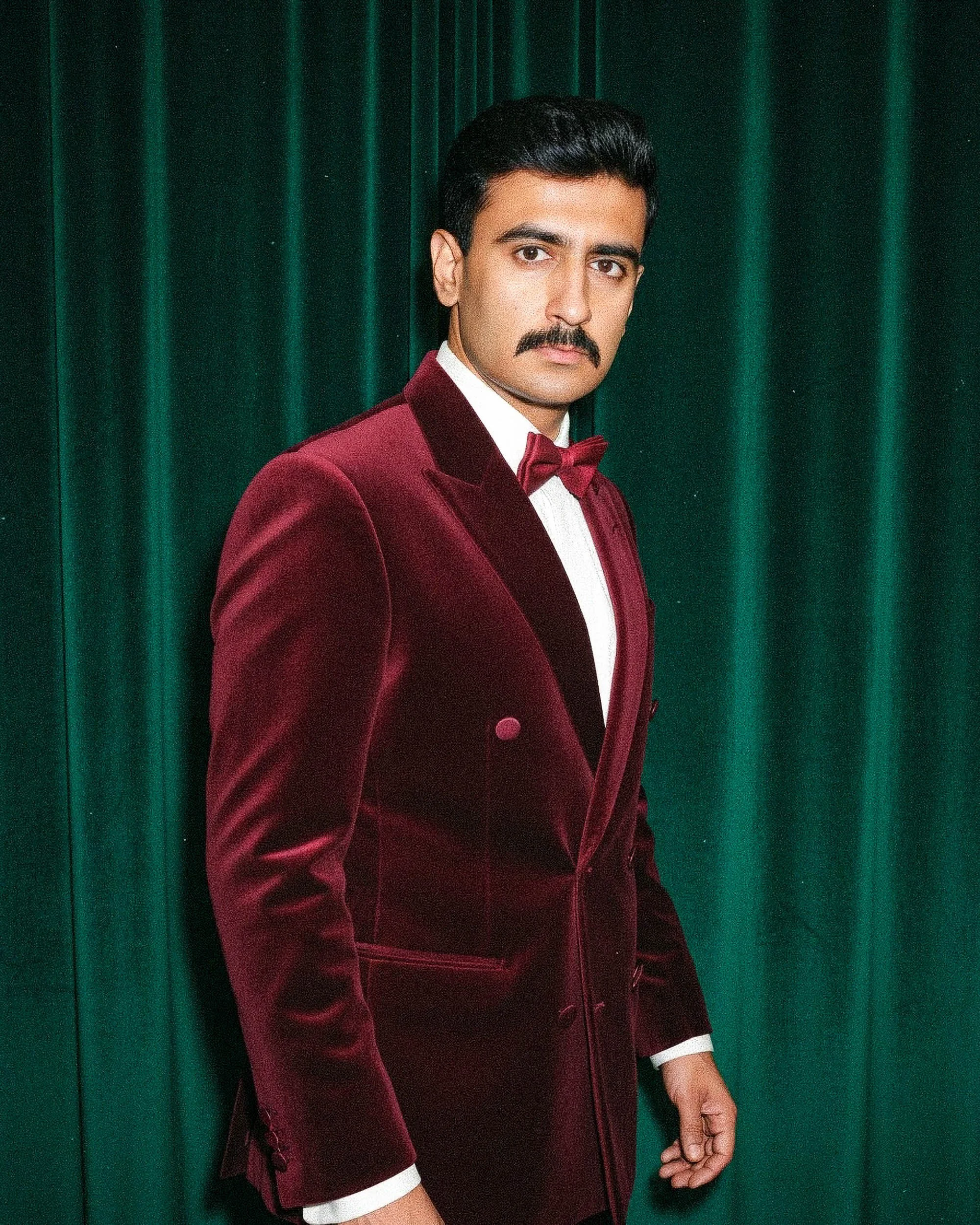 A man with a dark mustache and dark hair wearing a white shirt, maroon velvet tuxedo with a matching bow tie, standing in front of dark green curtains.