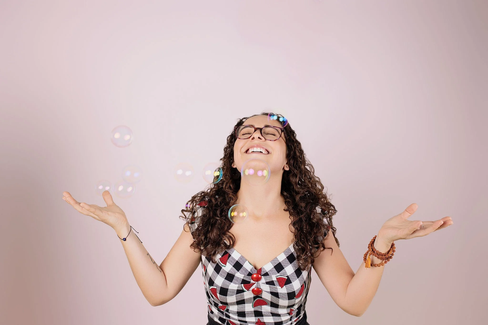 Pessoa sorrindo com os braços abertos, usando óculos, brincando com bolhas de sabão, vestida com vestido de padrão xadrez com fatias de melancia