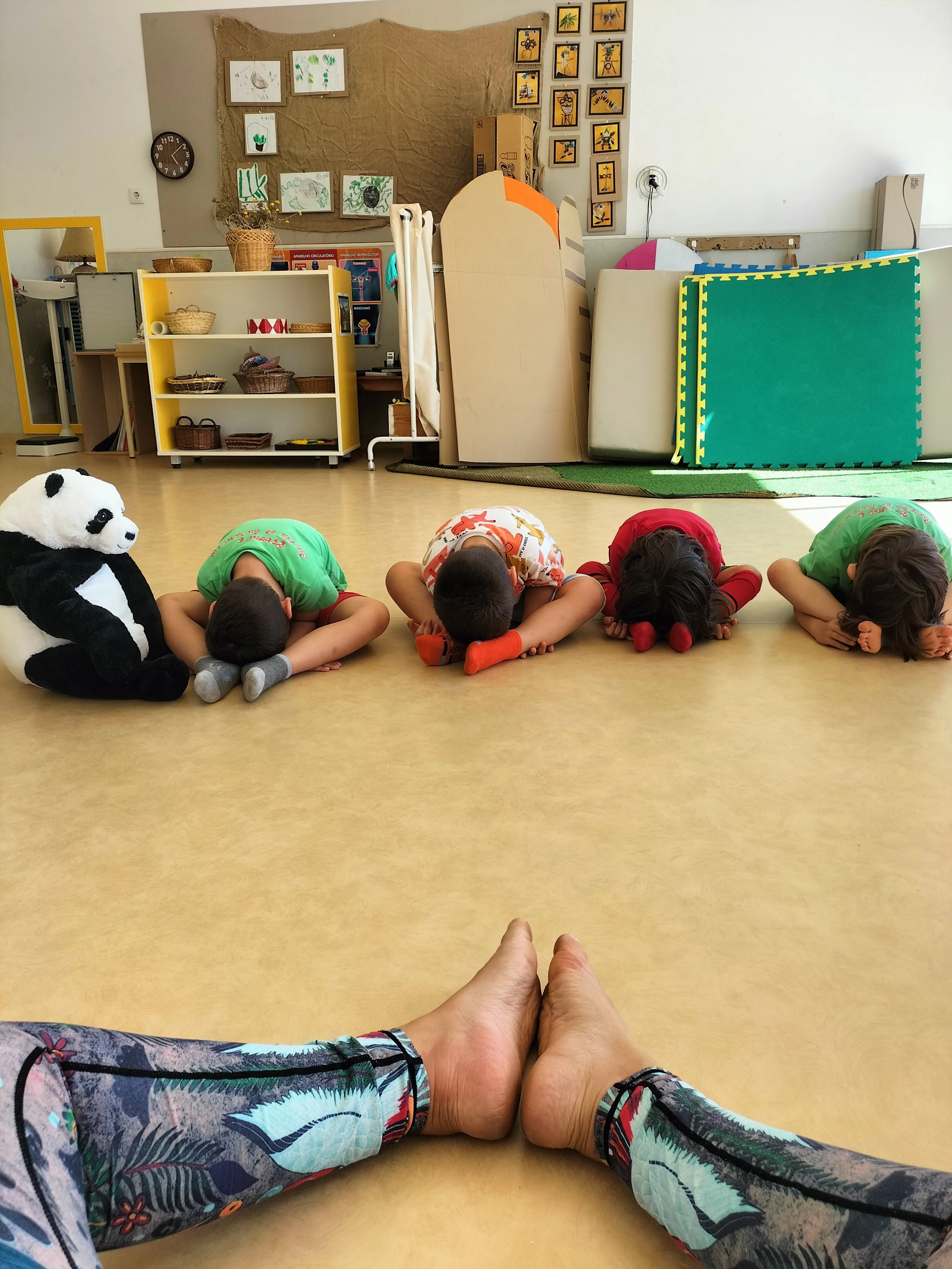Imagem de uma sala de aula, com crianças sentado no chão com cabeça abaixada e mãos cobrindo o rosto. Uma boneca de panda gigante está ao lado de uma criança. No fundo há quadros na parede e móveis de armazenamento.