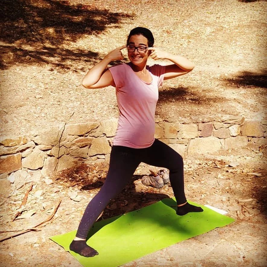 Mulher praticando yoga ao ar livre, fazendo uma postura com uma perna dobrada e os braços ajustados na cabeça, em uma esteira verde e fundo de pedra e chão de terra.