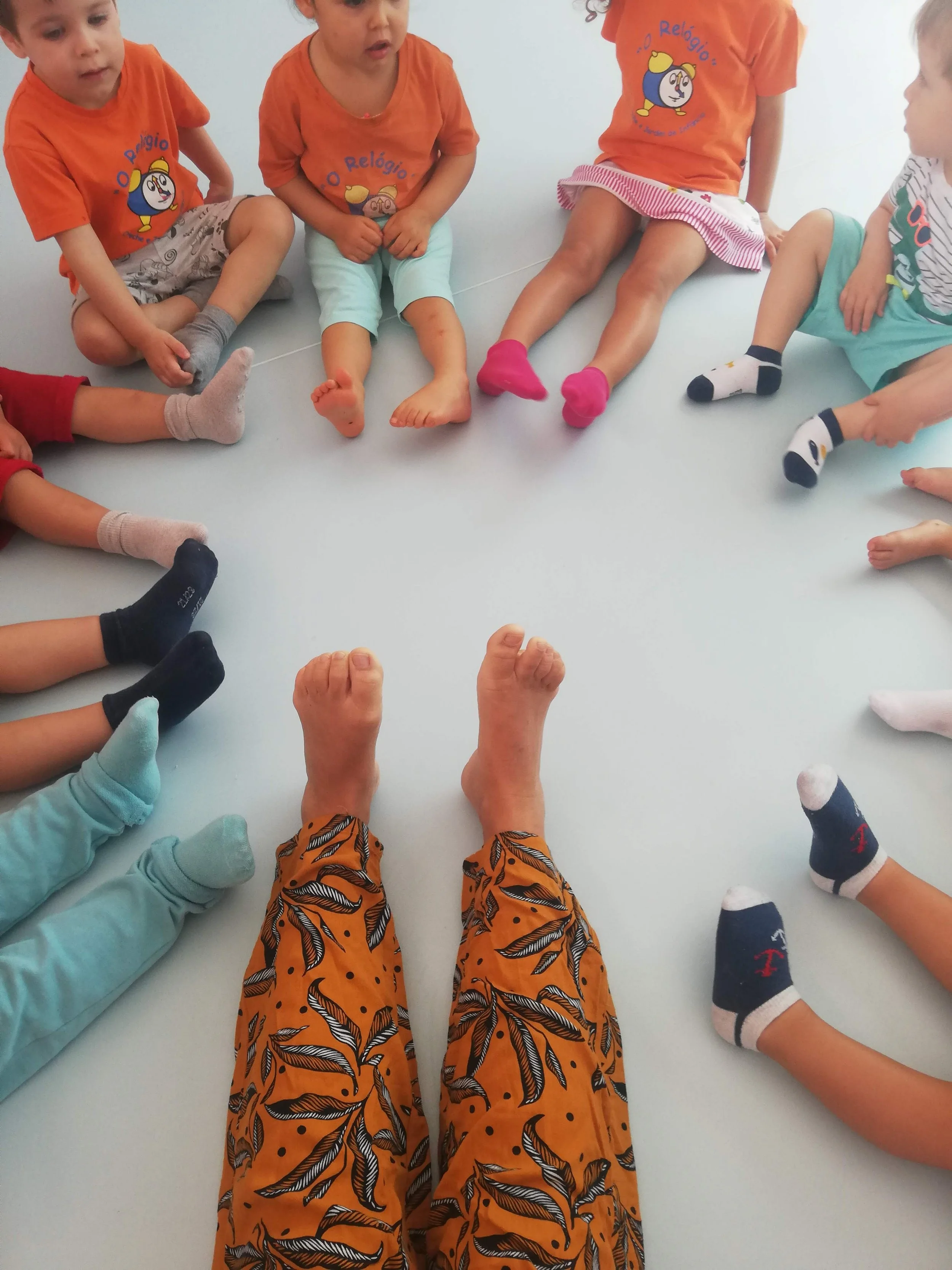 Crianças vestidas com roupas de cores variadas e meias diferentes, sentadas em círculo no chão, com as pernas esticadas à frente.