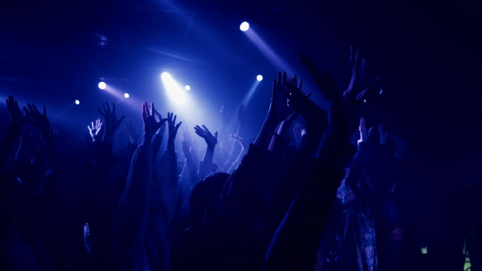 People raising their hands at a concert or dance event with blue lighting and spotlight effects.