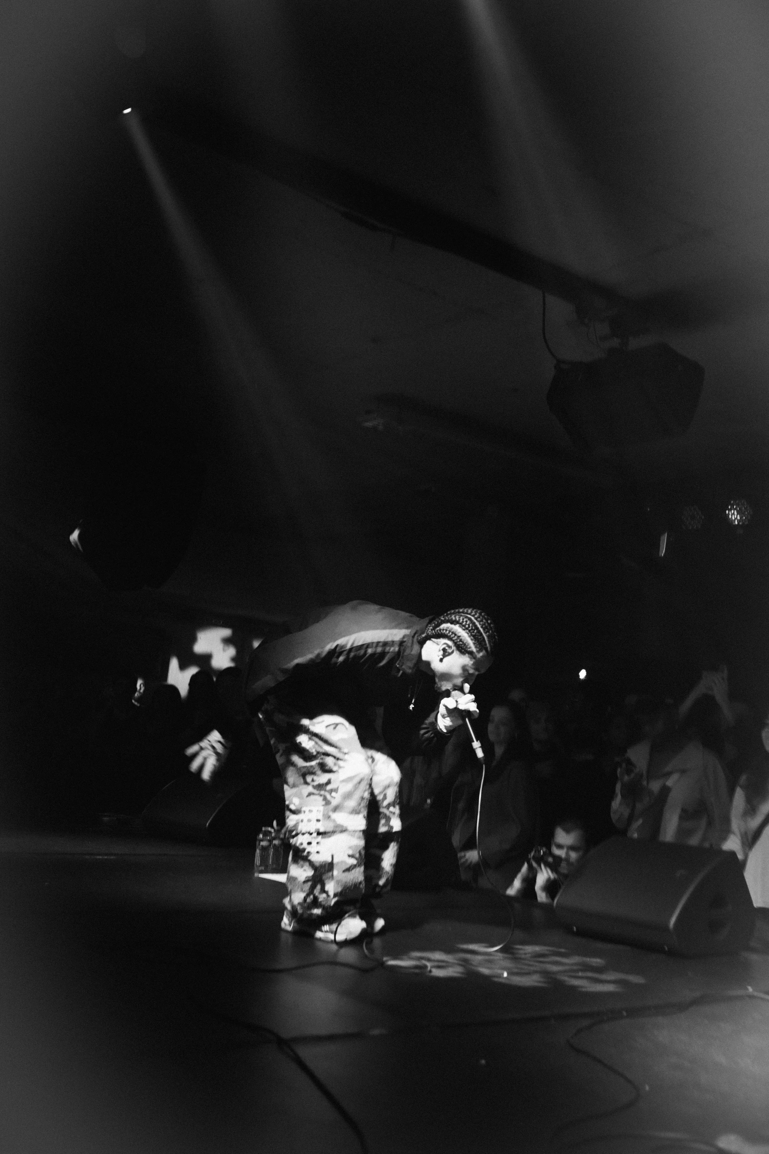 A performer with camo pants and a striped winter hat singing into a microphone on stage, with an audience watching.