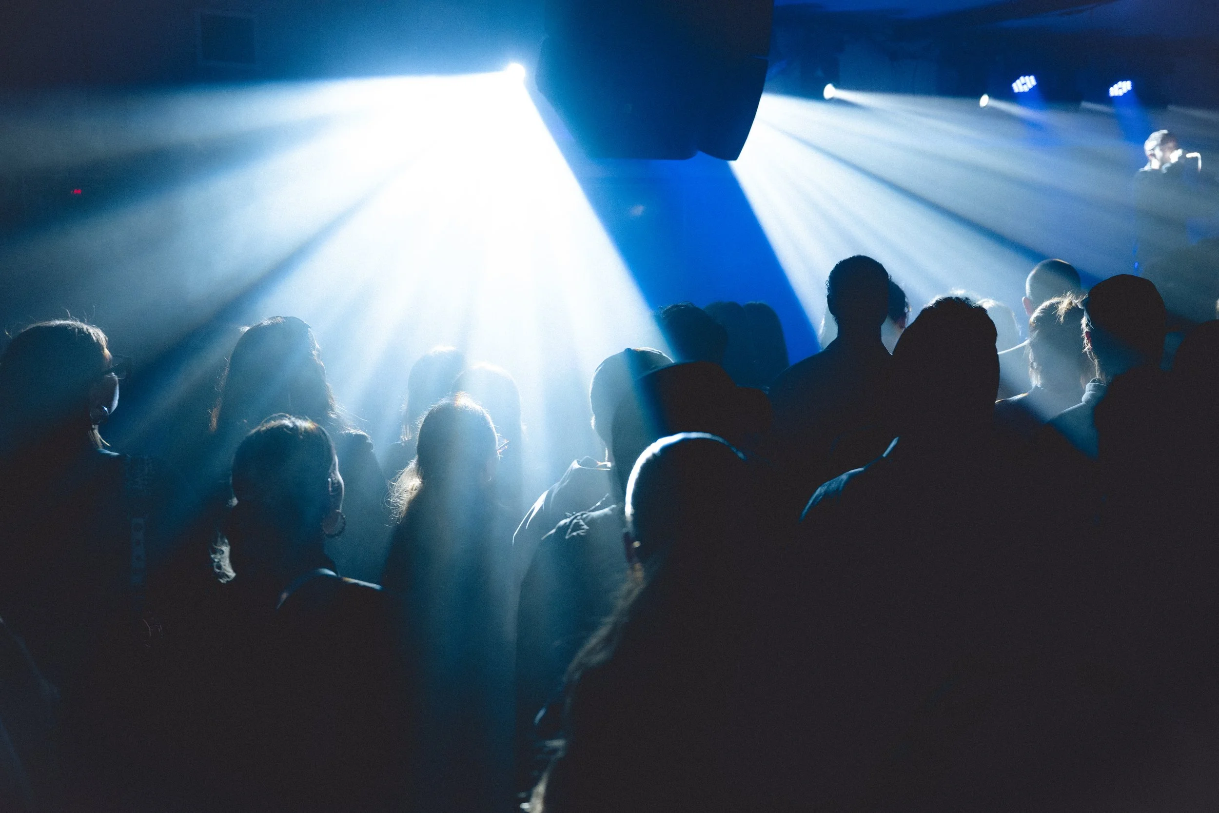 People at a concert or nightclub with bright blue and white stage lights creating beams through the dark venue.