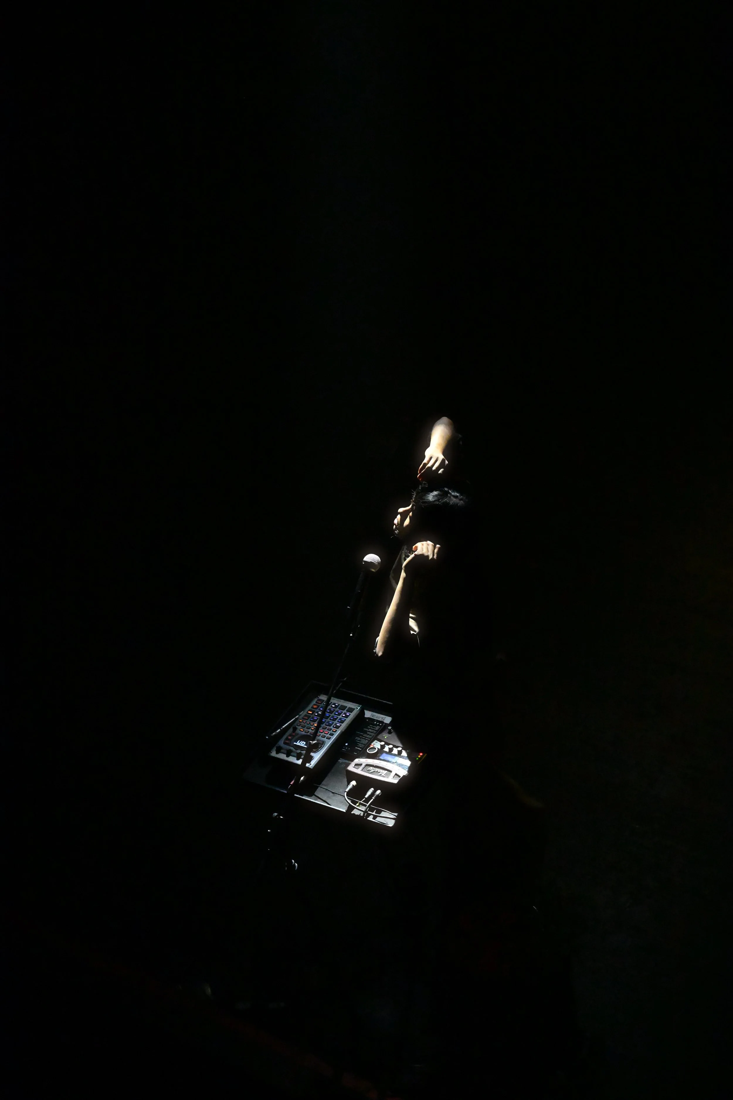 A person performing on stage, illuminated by a single spotlight, with DJ equipment in front of them.