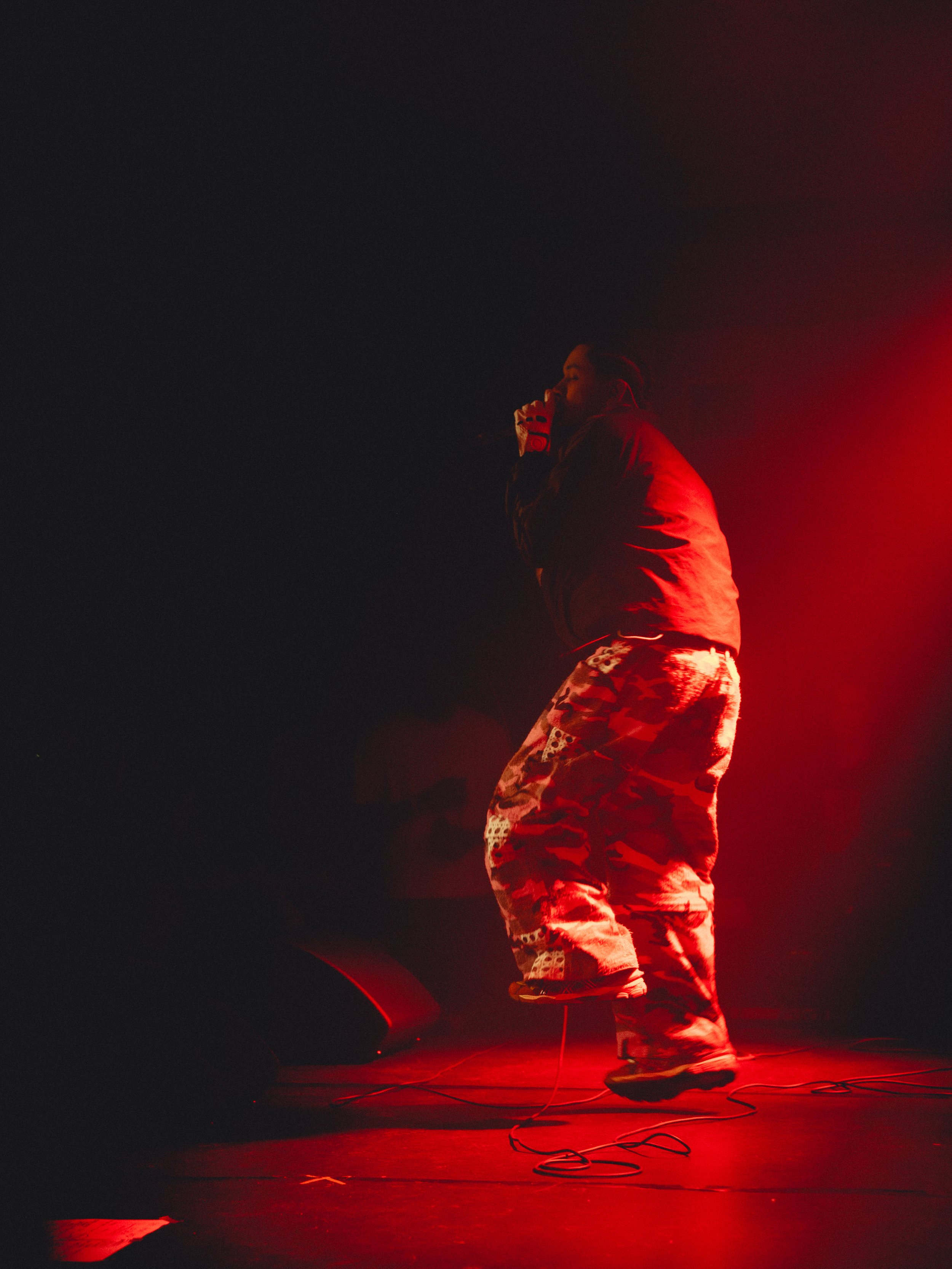 A person standing on stage in front of a dark background, illuminated by red stage lighting, wearing camouflage pants, a jacket, and holding a microphone.