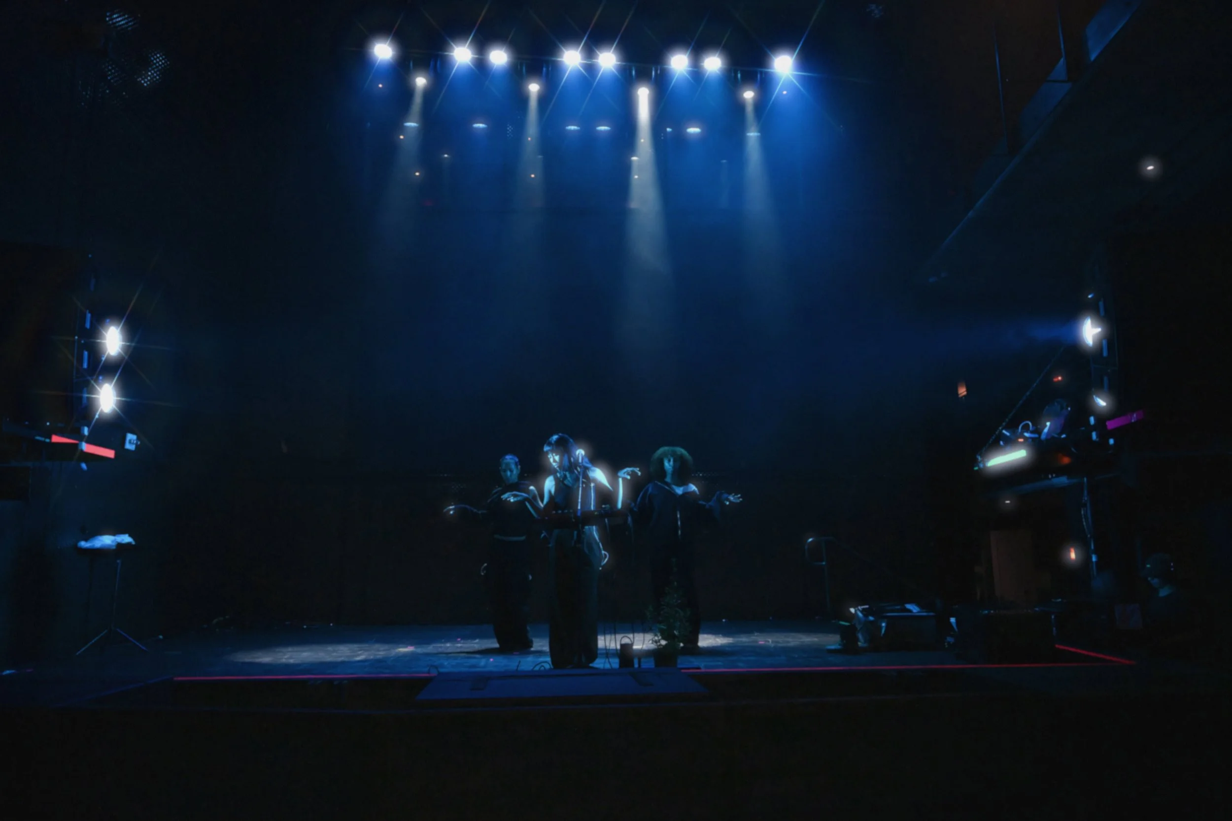 Dark stage with blue lighting and five performers standing close together, possibly preparing for a performance or rehearsal.