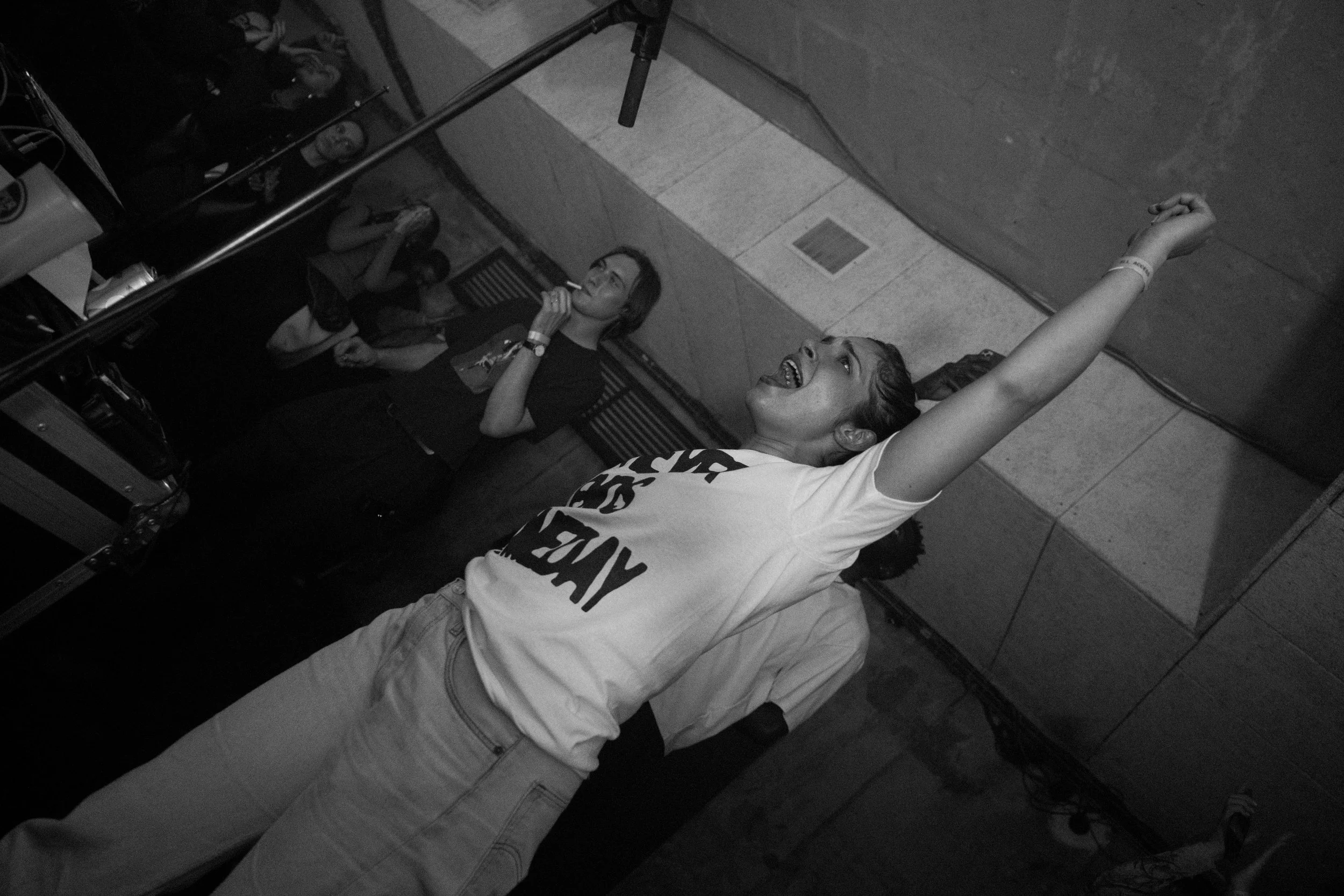 A woman in a white T-shirt with black lettering, gray pants, and an identification wristband on her right wrist, standing with her arm raised and smiling, in front of a mirror in a room with tiled walls. Two other women are visible in the background;