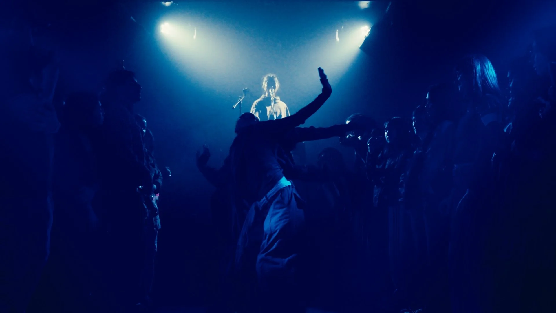 A person dancing in front of a small audience in a dark club, illuminated by blue and white stage lights, with a vocalist performing on stage in the background.