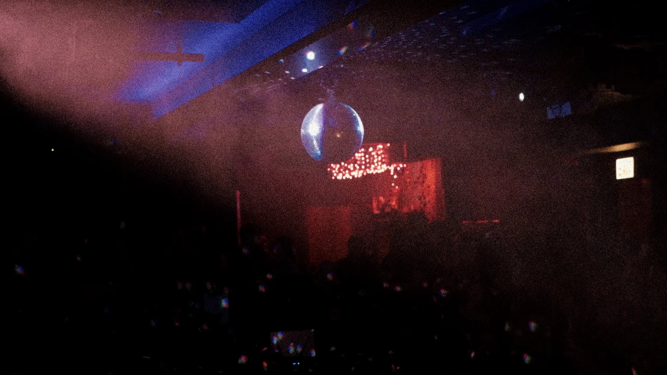 A dark nightclub scene with colorful lights, a disco ball hanging from the ceiling, and a musicians on stage in the background.