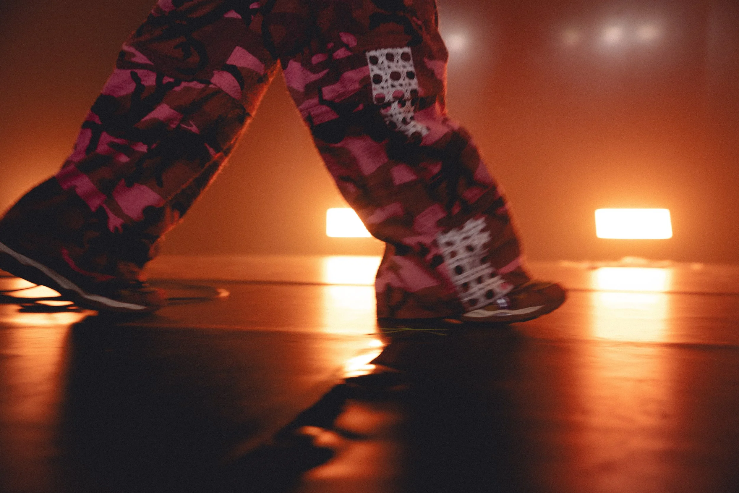 Close-up of a person riding a skateboard indoors, with blurred background lights and reflections on the floor, wearing patterned clothing.