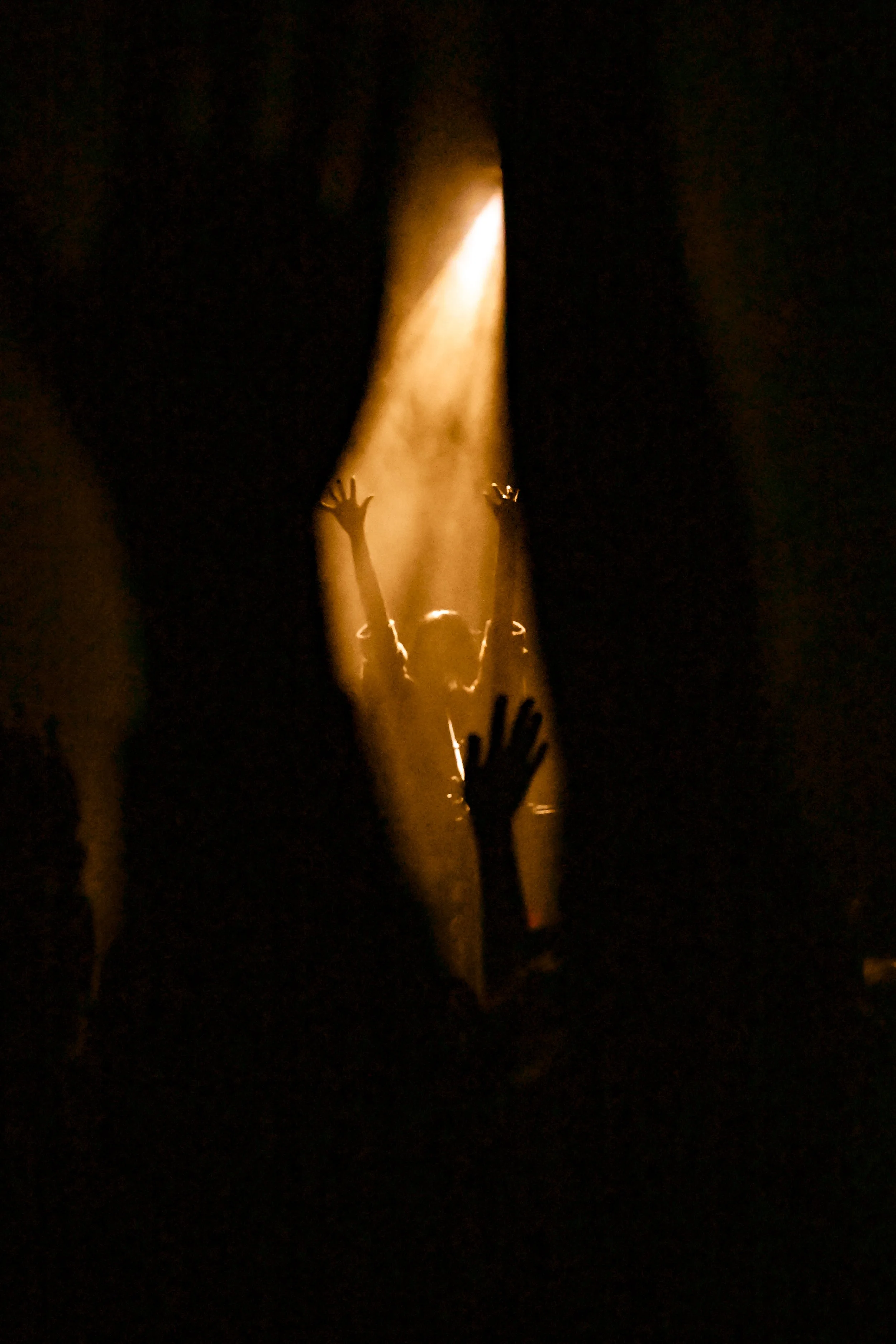 A dark scene with a bright yellow spotlight shining down on a scene with hands reaching upward, possibly at a concert or performance.
