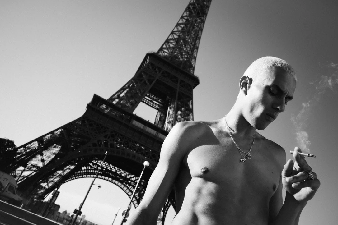 A shirtless man with a shaved head holding a cigarette near the Eiffel Tower in Paris, France.