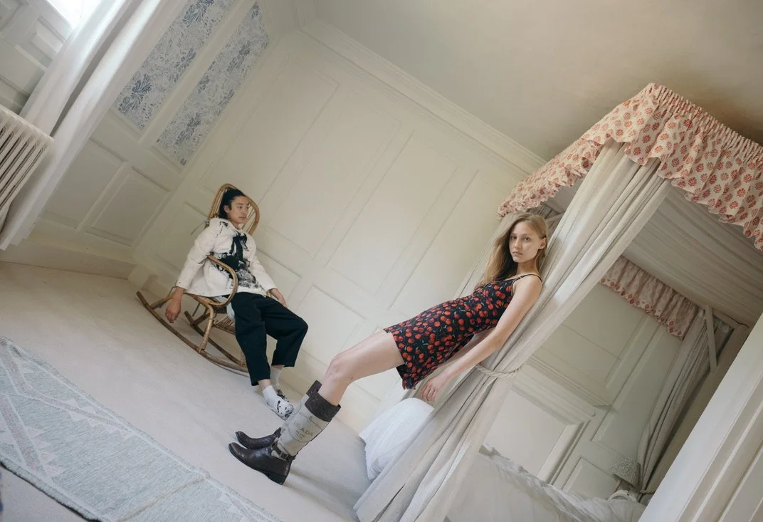 Two women in a vintage room with white walls. One woman with blonde hair sits on the edge of a bed, wearing a black floral dress and dark boots. The other woman with dark hair, dressed in a white jacket and black pants, sits on a wooden rocking chair