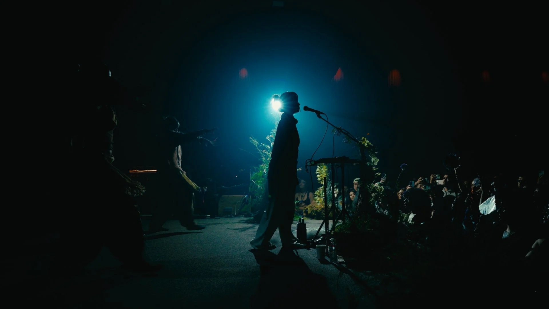 A silhouette of a performer singing into a microphone at an indoor concert with audience members capturing the moment on their phones, illuminated by a blue backlight.