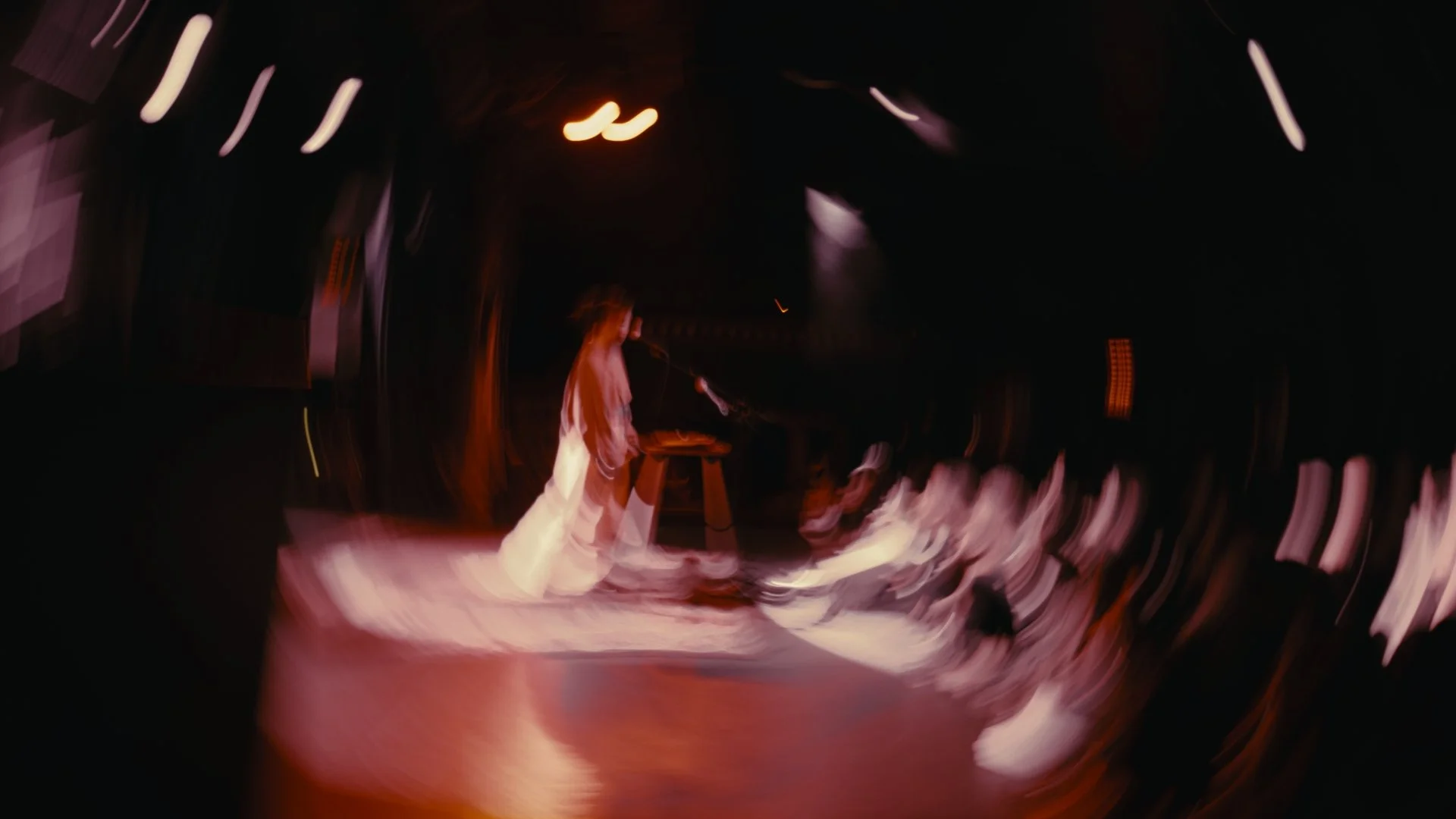 A person stands on stage in front of a microphone, wearing light-colored clothing, with a piano nearby, in a darkened room with blurred audience and stage lights.