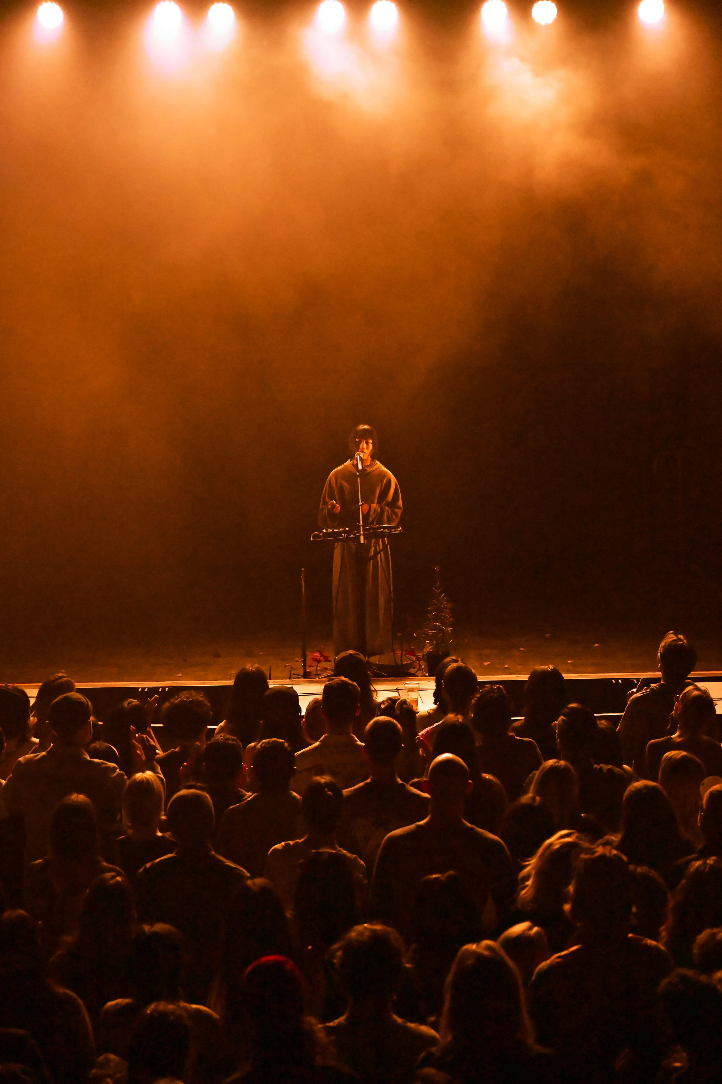 A performer on stage, wearing a brown hoodie, stands in front of a microphone and keyboard. The stage is lit with warm, orange spotlights, and there is an audience seated in front.