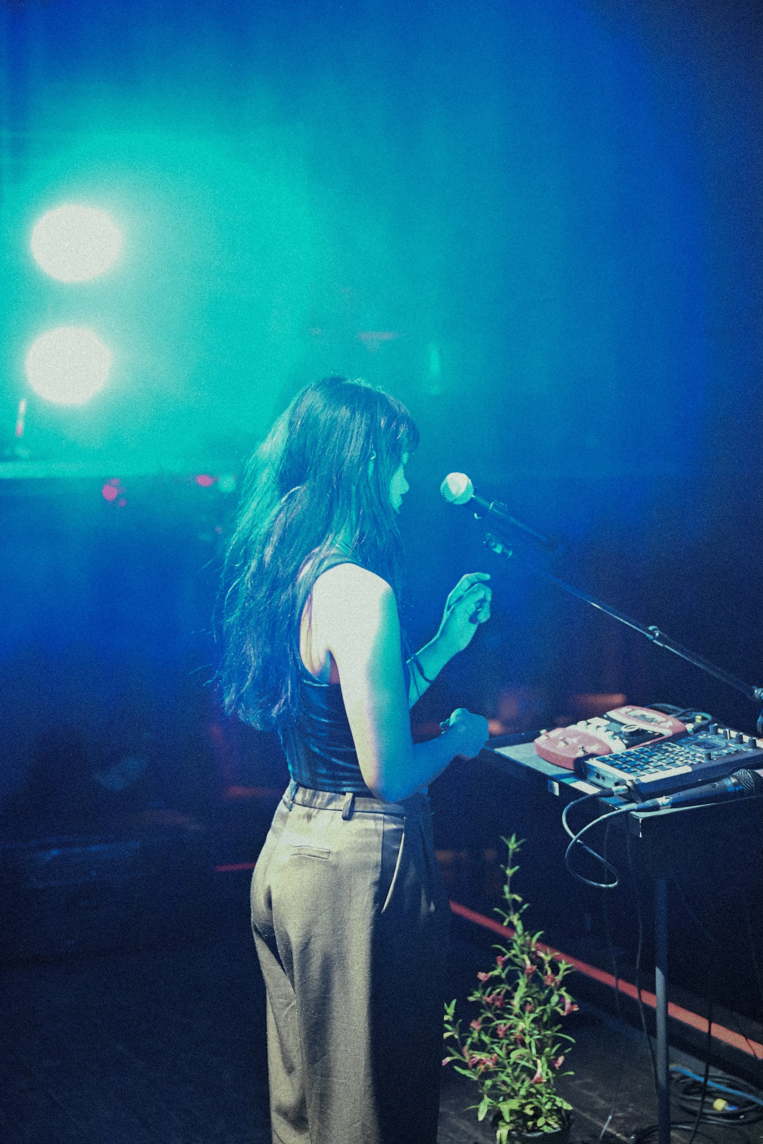 A woman with long hair, wearing a sleeveless top and high-waisted pants, standing at a DJ setup with turntables and a mixer, in a dimly lit environment with stage lights in the background.
