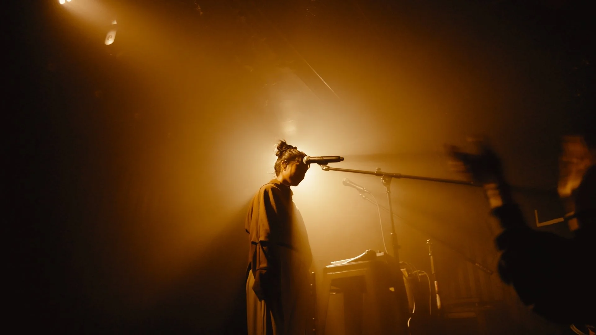 A person stands at a microphone on stage, illuminated by warm, yellowish stage lighting, with their hair tied up in a bun and wearing a brown jacket, while performing in a dark venue.
