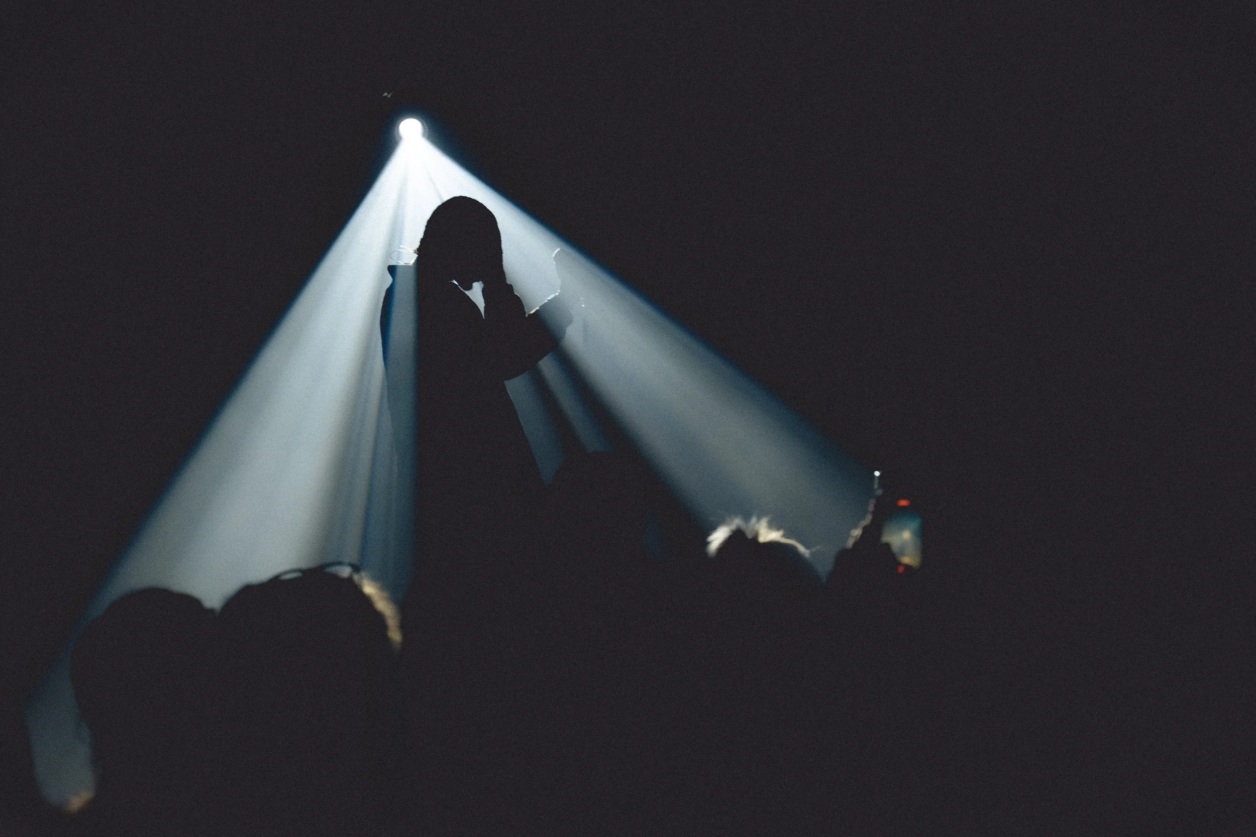 Dark background with a small bright light source at the top illuminating a person's silhouette with long hair, standing among other figures and objects, possibly at an event or performance with dramatic lighting.