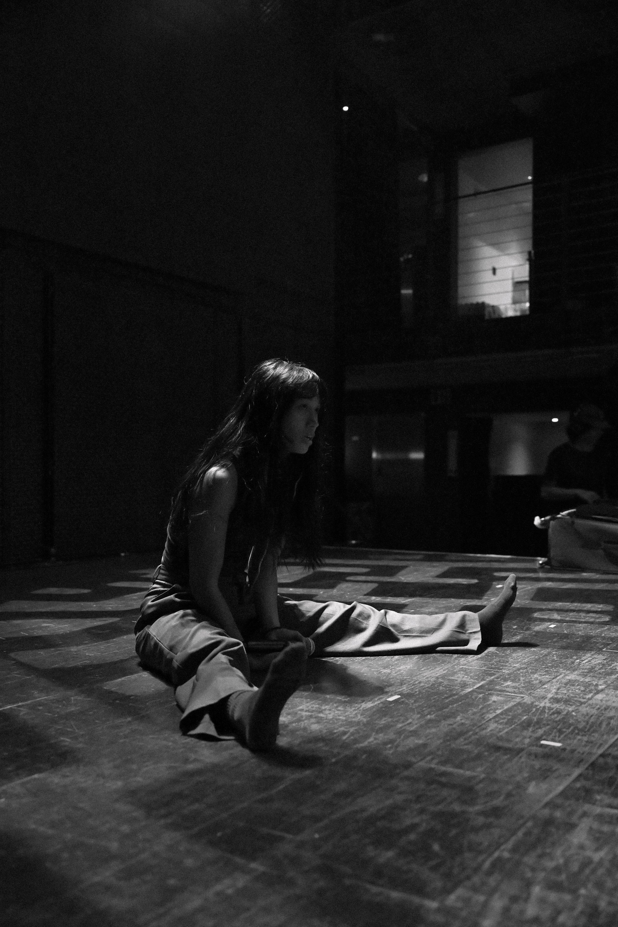 A woman practicing yoga on a dimly lit wooden floor, sitting on the floor with one leg extended and the other bent, her hands resting on her bent knee, with a background of dark walls and a window showing a night sky.