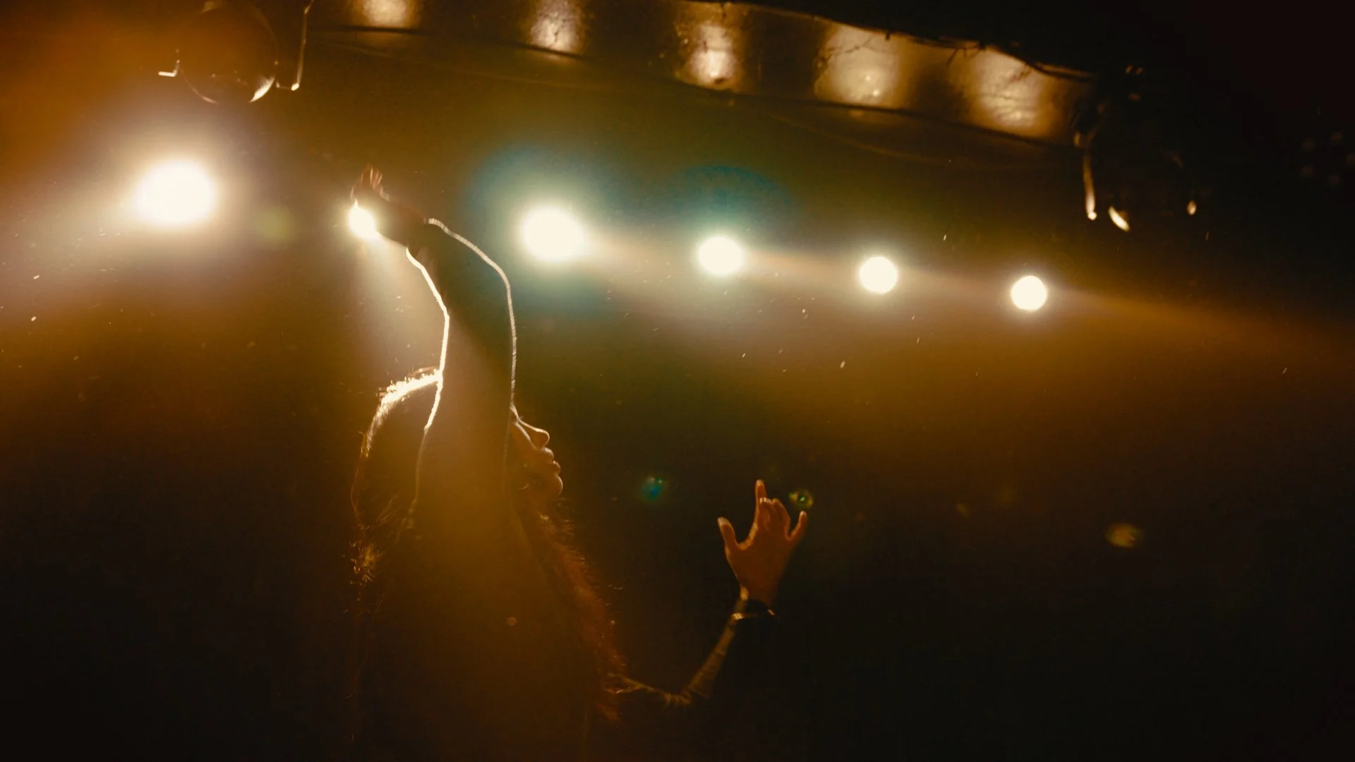 A person with long hair and a bracelet on their wrist is raising their hand with fingers extended towards a row of bright stage lights, creating a starry effect in a dark environment.
