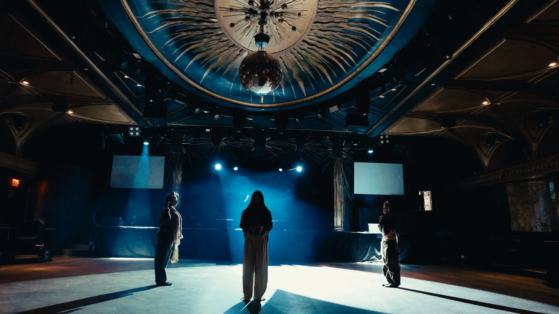 A dimly lit dance studio or event space with a shiny disco ball hanging from a decorative ceiling. Three people stand on the floor, shadowed by blue stage lights, with large screens or windows on the walls.