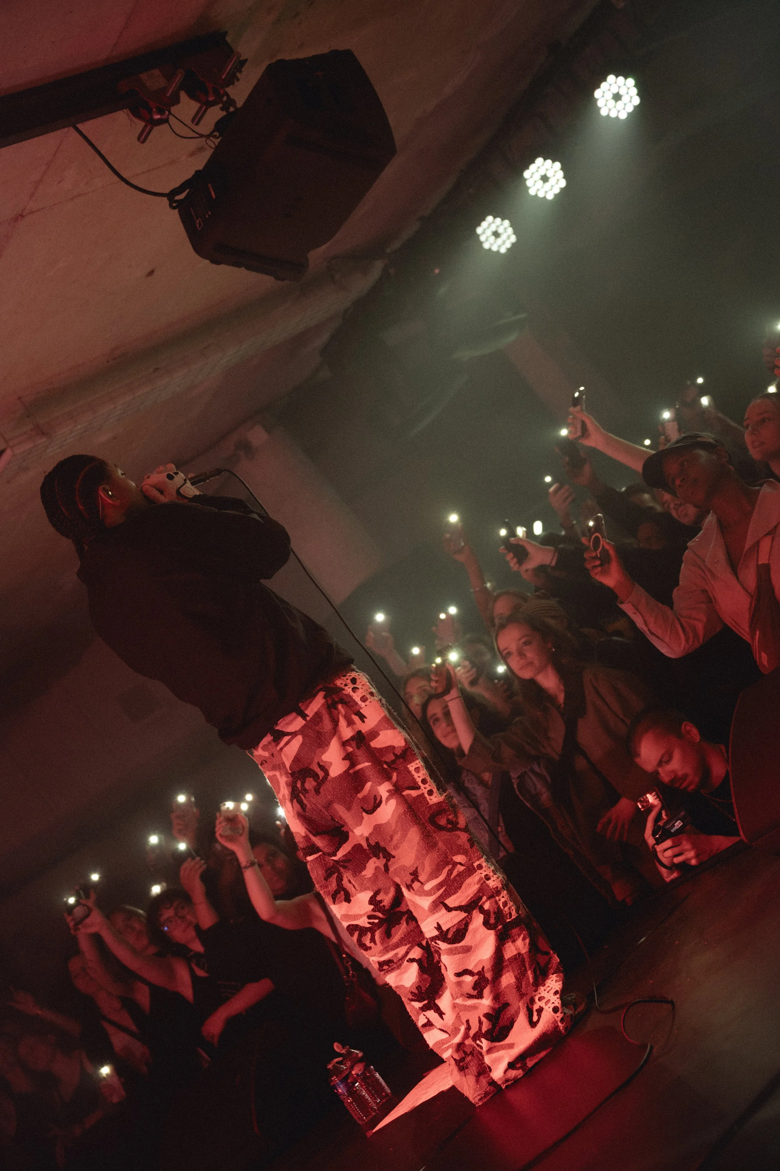 A woman stands on stage with a microphone in front of a large crowd. Audience members are holding up smartphones with lights on, creating a glow. The scene is dimly lit with red lighting. The woman is wearing patterned pants and a dark top.